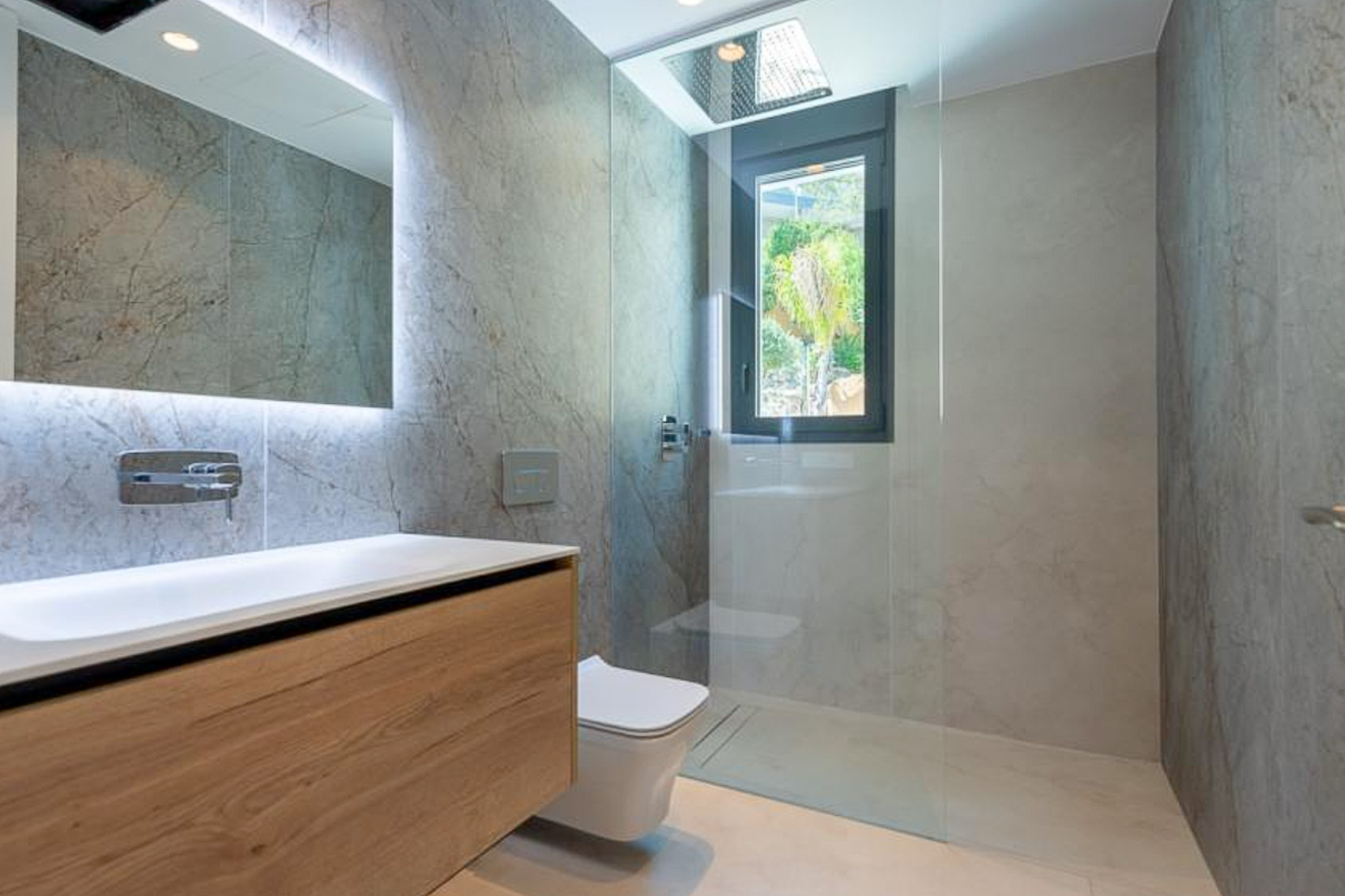 Baño de diseño con revestimiento efecto piedra, mueble de madera suspendido, espejo retroiluminado y ducha con rociador empotrado en el techo.