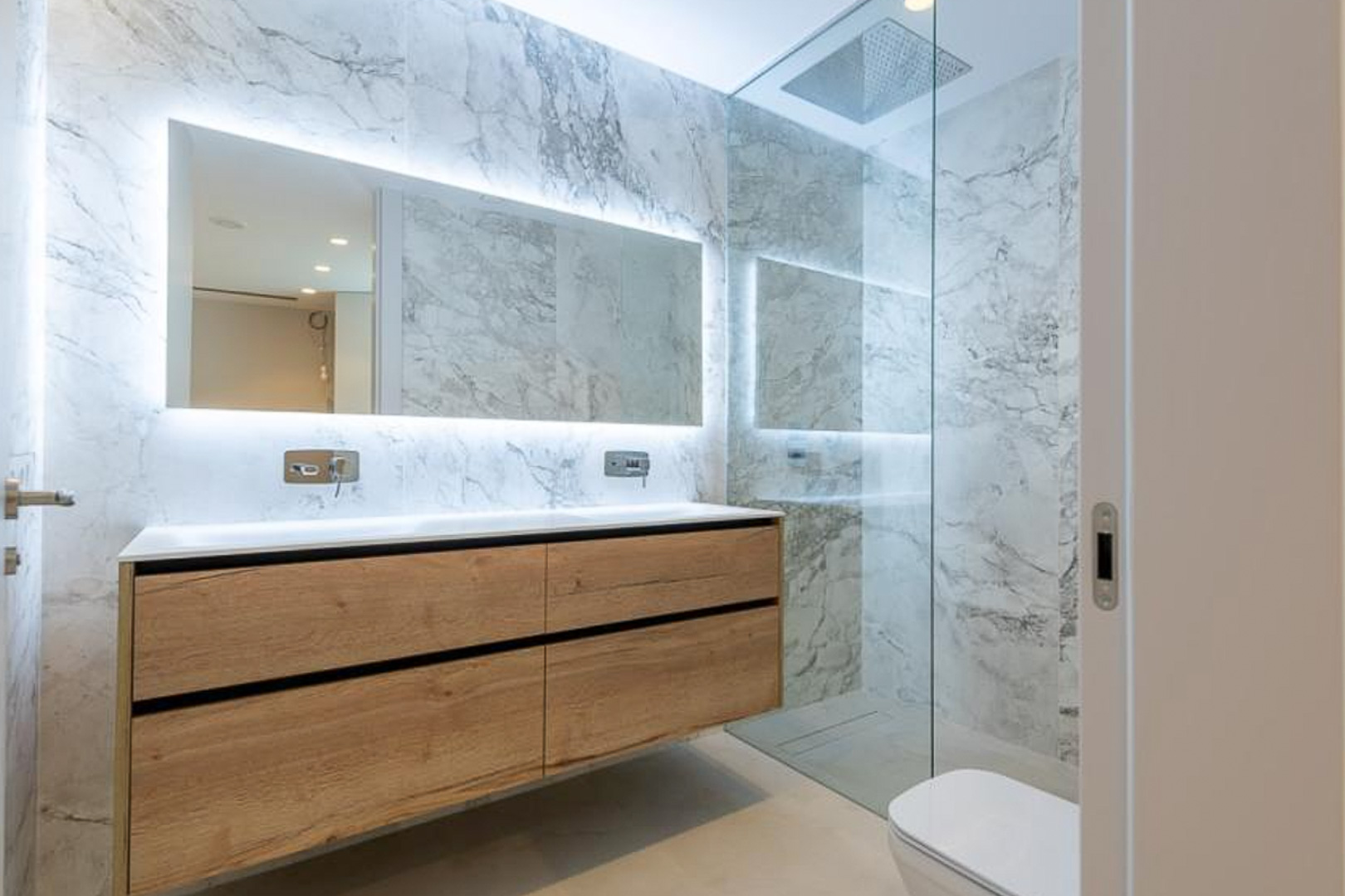 Baño moderno con revestimiento de mármol, mueble suspendido de madera con doble lavabo y espejo retroiluminado. Incluye ducha de obra con mampara de cristal.