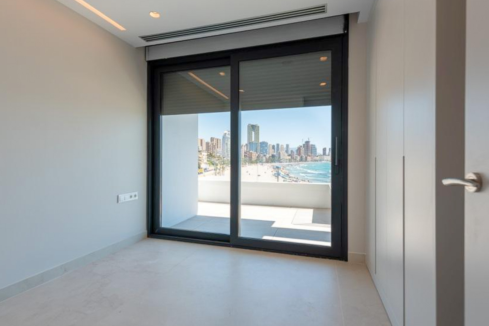 Dormitorio con armarios empotrados de suelo a techo y acceso directo a terraza con vistas panorámicas a la playa y el skyline de Benidorm.