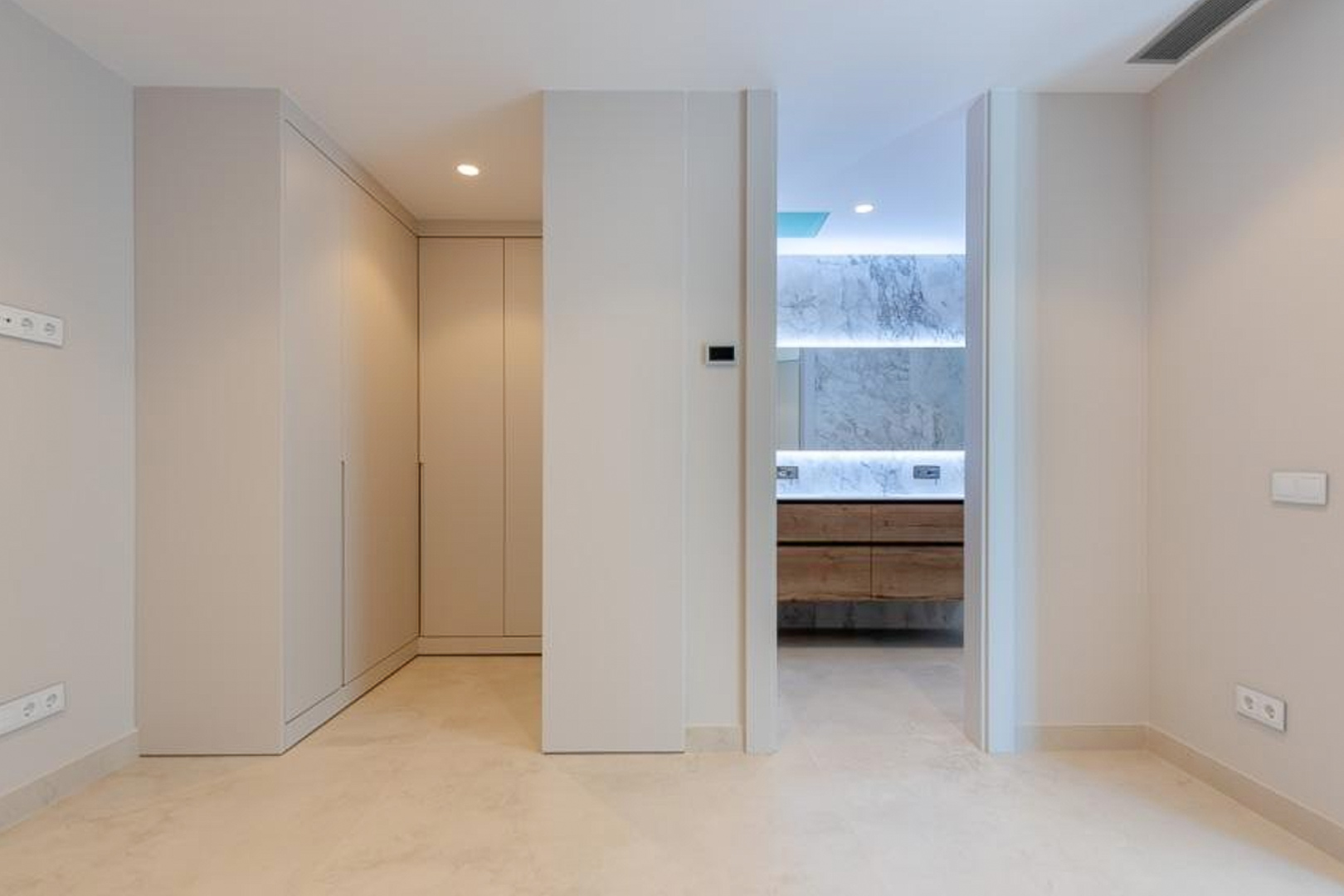 Dormitorio principal con armarios empotrados de suelo a techo y acceso al baño en suite, con revestimiento de mármol, mobiliario de madera e iluminación LED integrada.