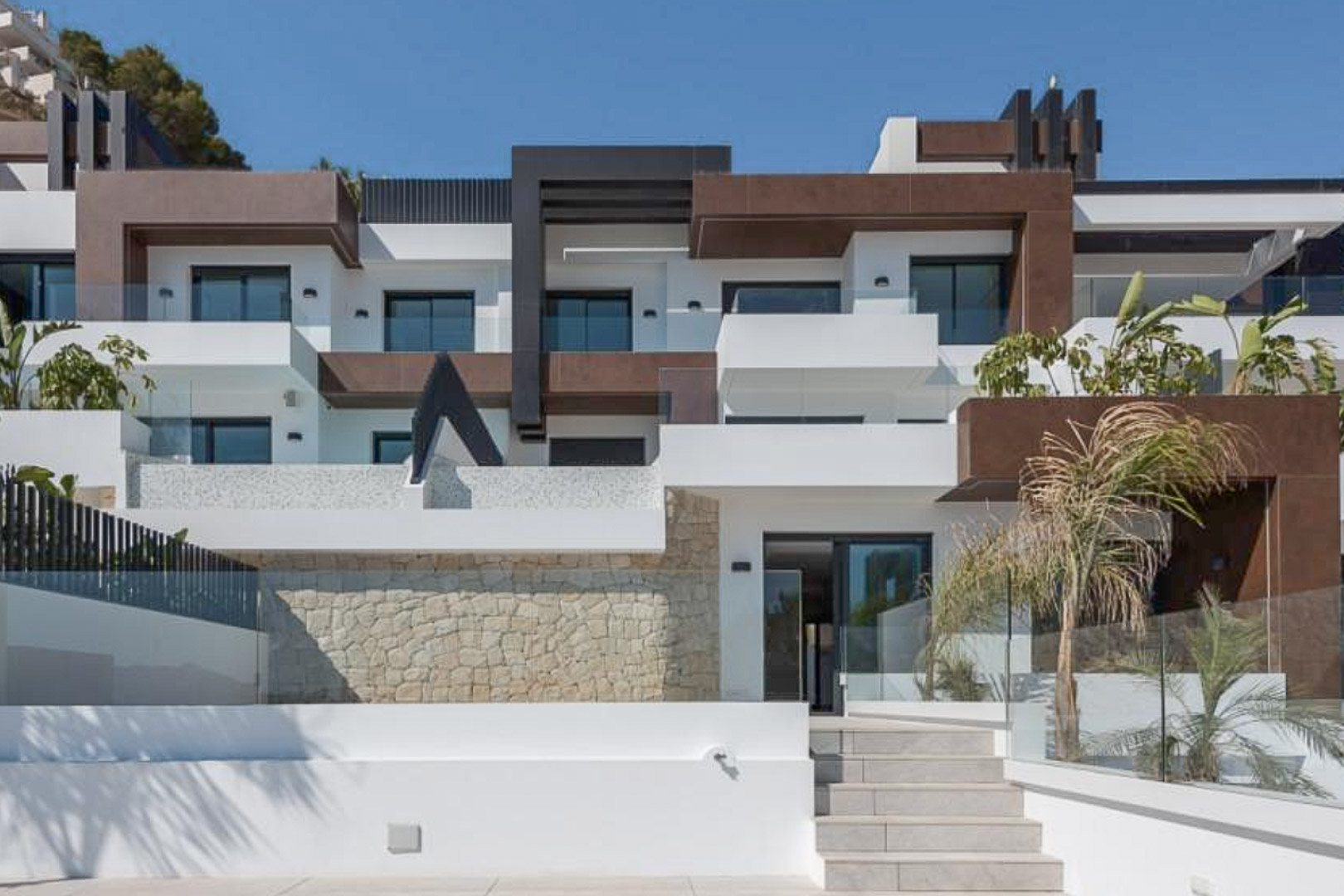 Fachada arquitectónica moderna con volúmenes en blanco y marrón, muros de piedra natural, barandillas de cristal y terrazas escalonadas bajo un cielo despejado.
