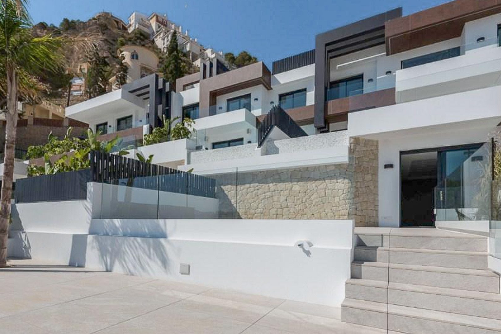 Fachada moderna de diseño escalonado con muros de piedra natural, amplias terrazas con barandillas de cristal y acceso mediante escalinata de gres porcelánico.