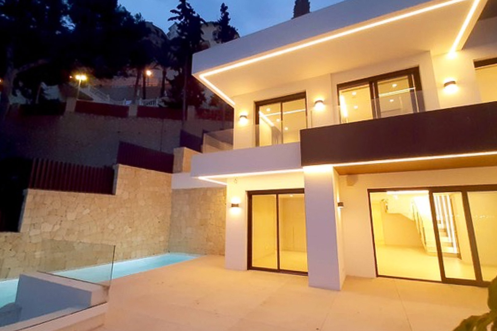 Fachada moderna de dos niveles con iluminación LED integrada, amplios ventanales correderos y terraza con piscina privada junto a un muro de piedra natural.