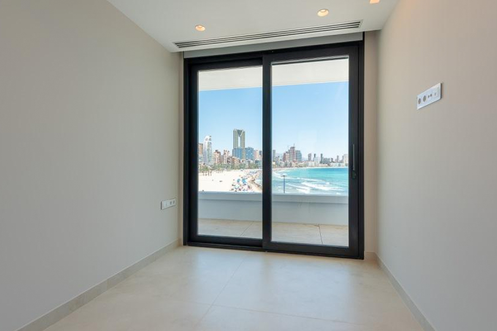 Habitación luminosa con suelo porcelánico claro y gran ventanal de corredera que ofrece vistas panorámicas a la playa y al skyline de Benidorm.