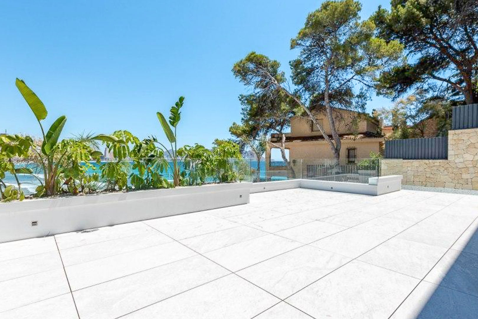 Terraza exterior con suelo de gres porcelánico de gran formato, barandilla de cristal sin marcos y jardineras integradas con plantas tropicales y vistas al mar.