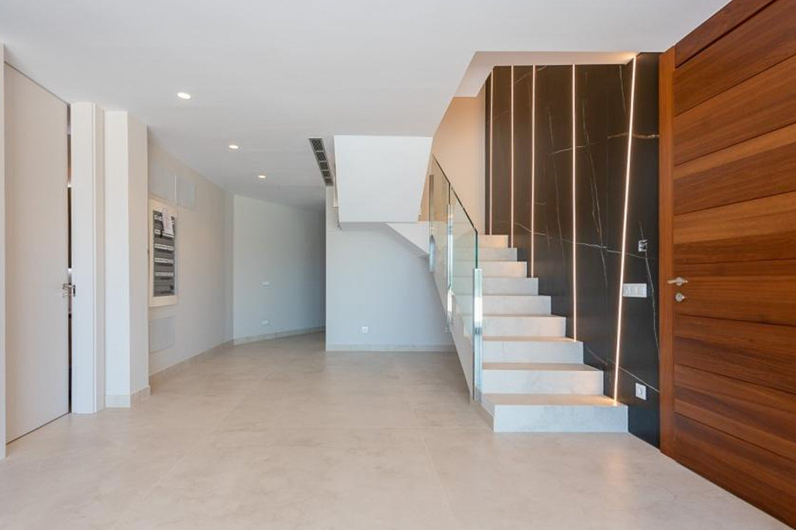 Vestíbulo de entrada con escalera de piedra blanca, barandilla de cristal y pared decorativa en mármol oscuro con tiras de iluminación LED integradas.