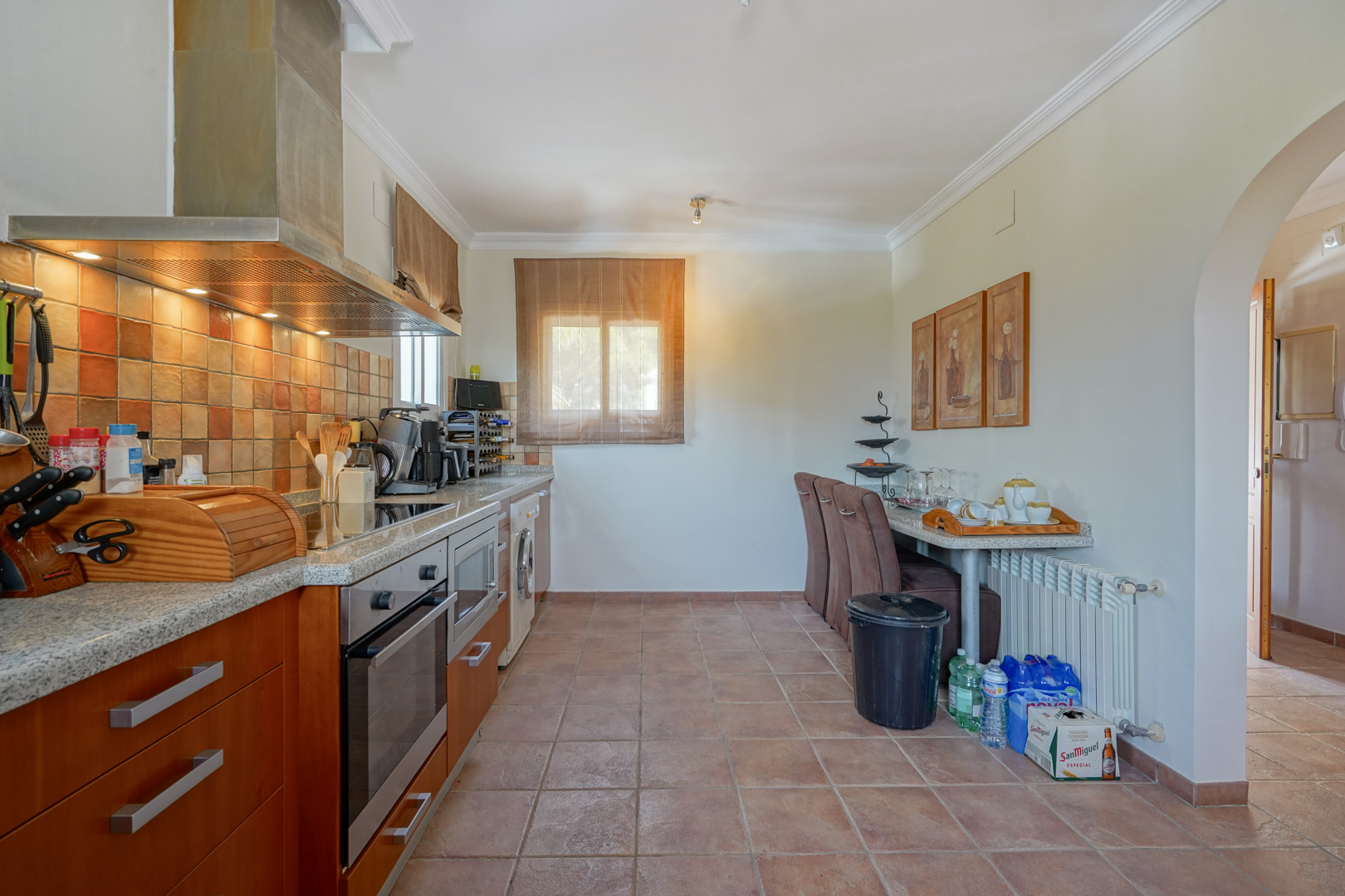 Cocina equipada con armarios de madera, encimeras de granito y electrodomésticos integrados, destacando los suelos de terracota y un acceso mediante arco tradicional.