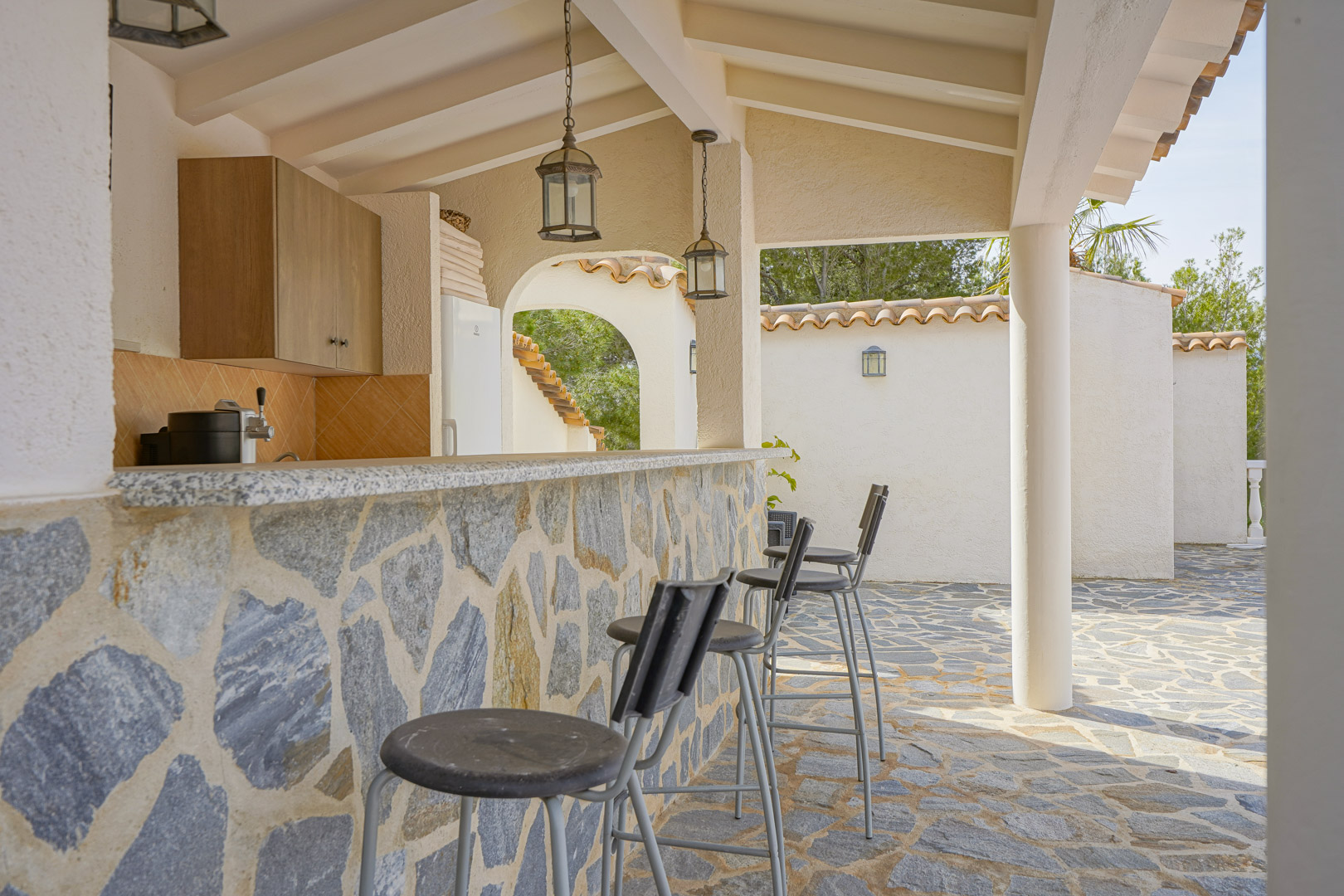 Cocina exterior con barra revestida en piedra natural, encimera de granito y taburetes altos bajo un porche con vigas de madera y lámparas colgantes de estilo rústico.