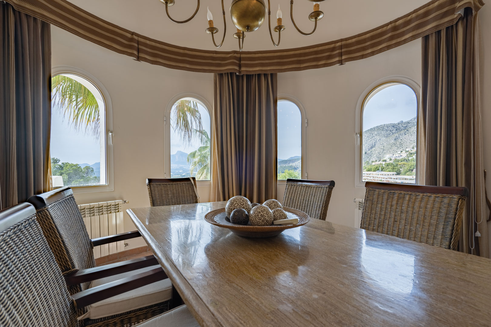 Comedor circular con mesa de piedra pulida y sillas de mimbre. Cuatro ventanas de arco ofrecen vistas panorámicas a la Sierra de Bernia y el paisaje montañoso de Altea.
