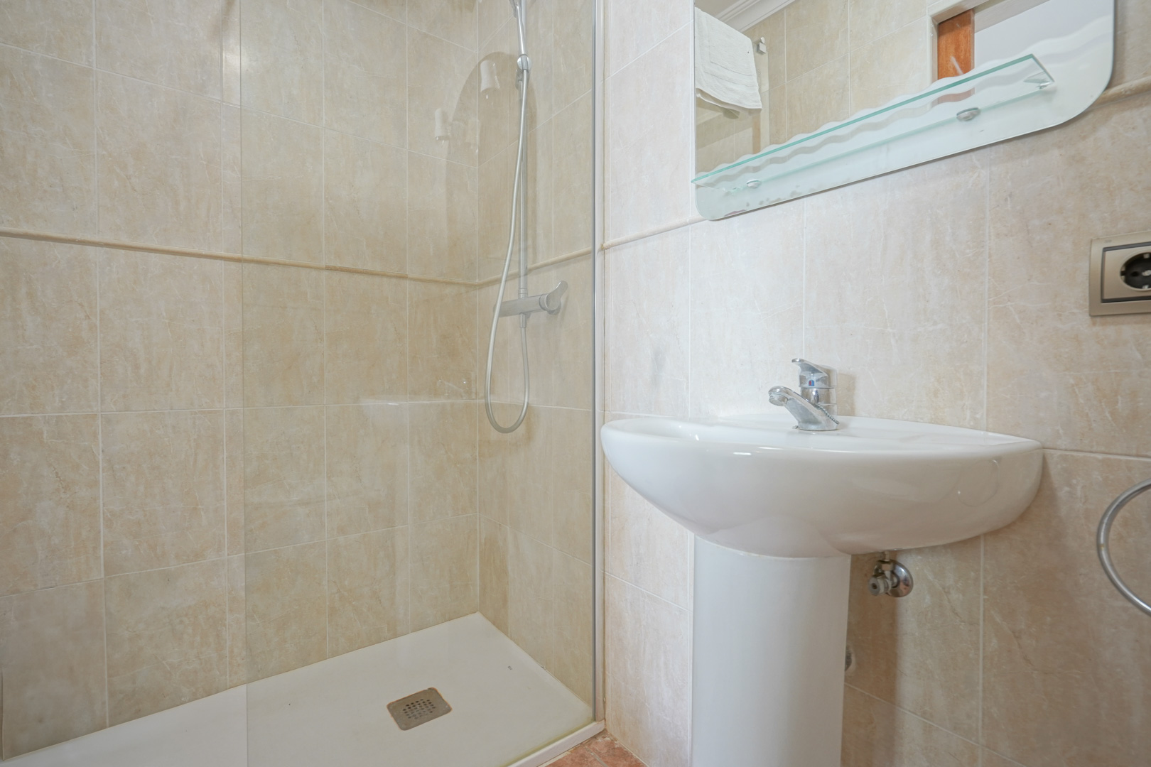 Cuarto de baño con azulejos en tonos beige, equipado con un lavabo de pedestal blanco y una ducha con mampara de cristal fija y grifería cromada.