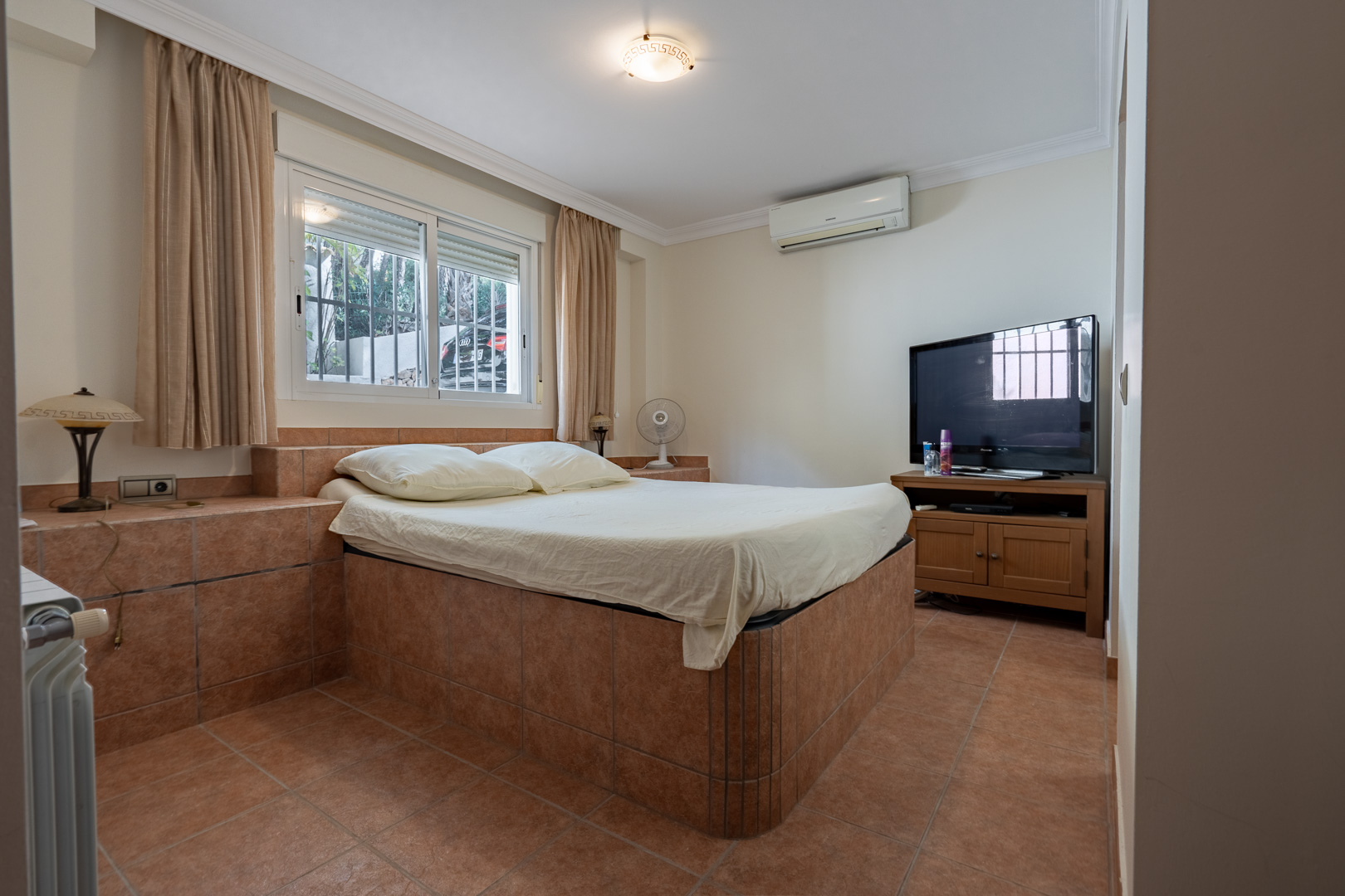 Dormitorio con base de cama integrada y suelos revestidos en gres cerámico, equipado con aire acondicionado, radiador y ventana exterior.