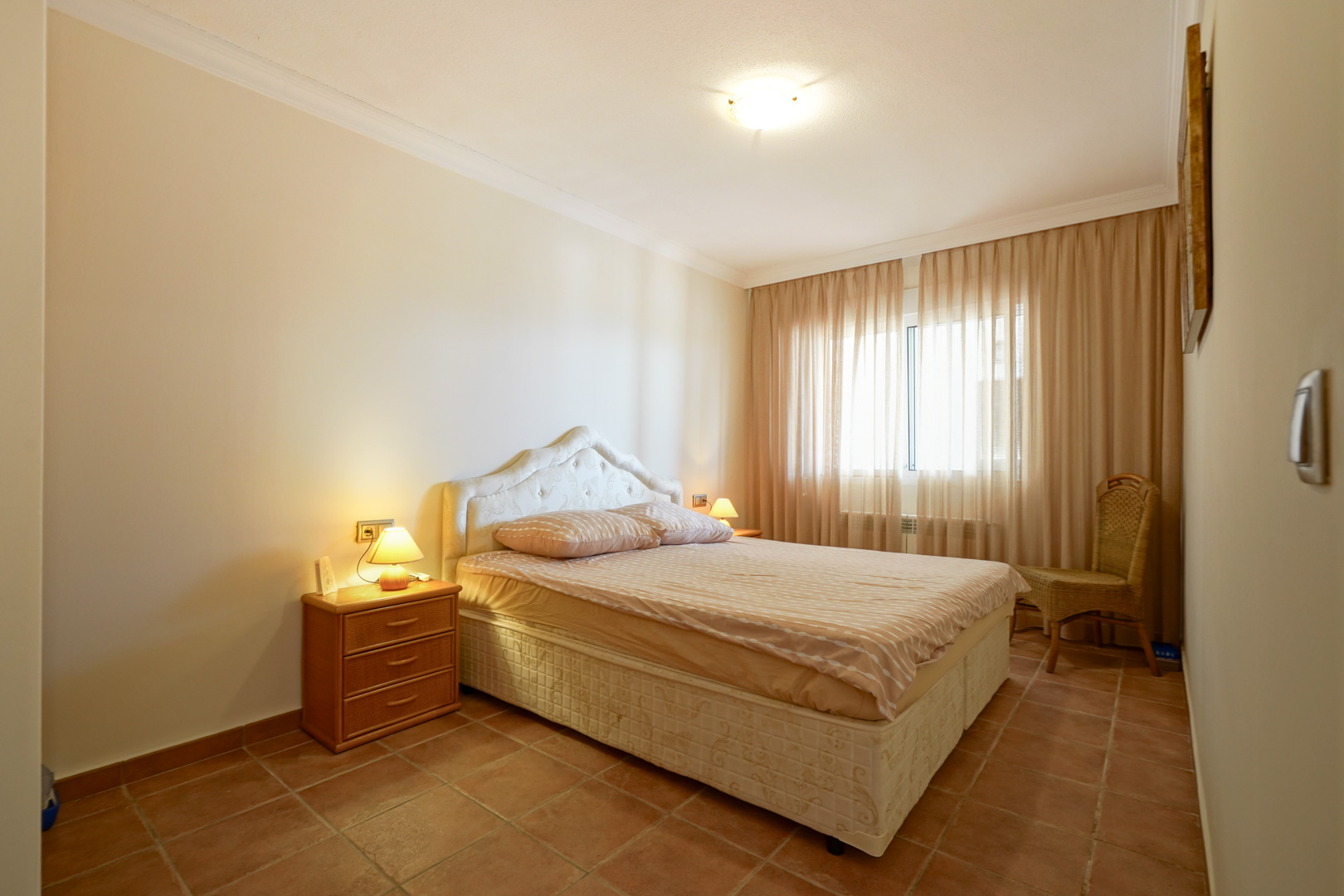 Dormitorio con suelos de baldosas de terracota, cama doble con cabecero tapizado, mesita de noche de madera y luz natural filtrada por cortinas translúcidas.