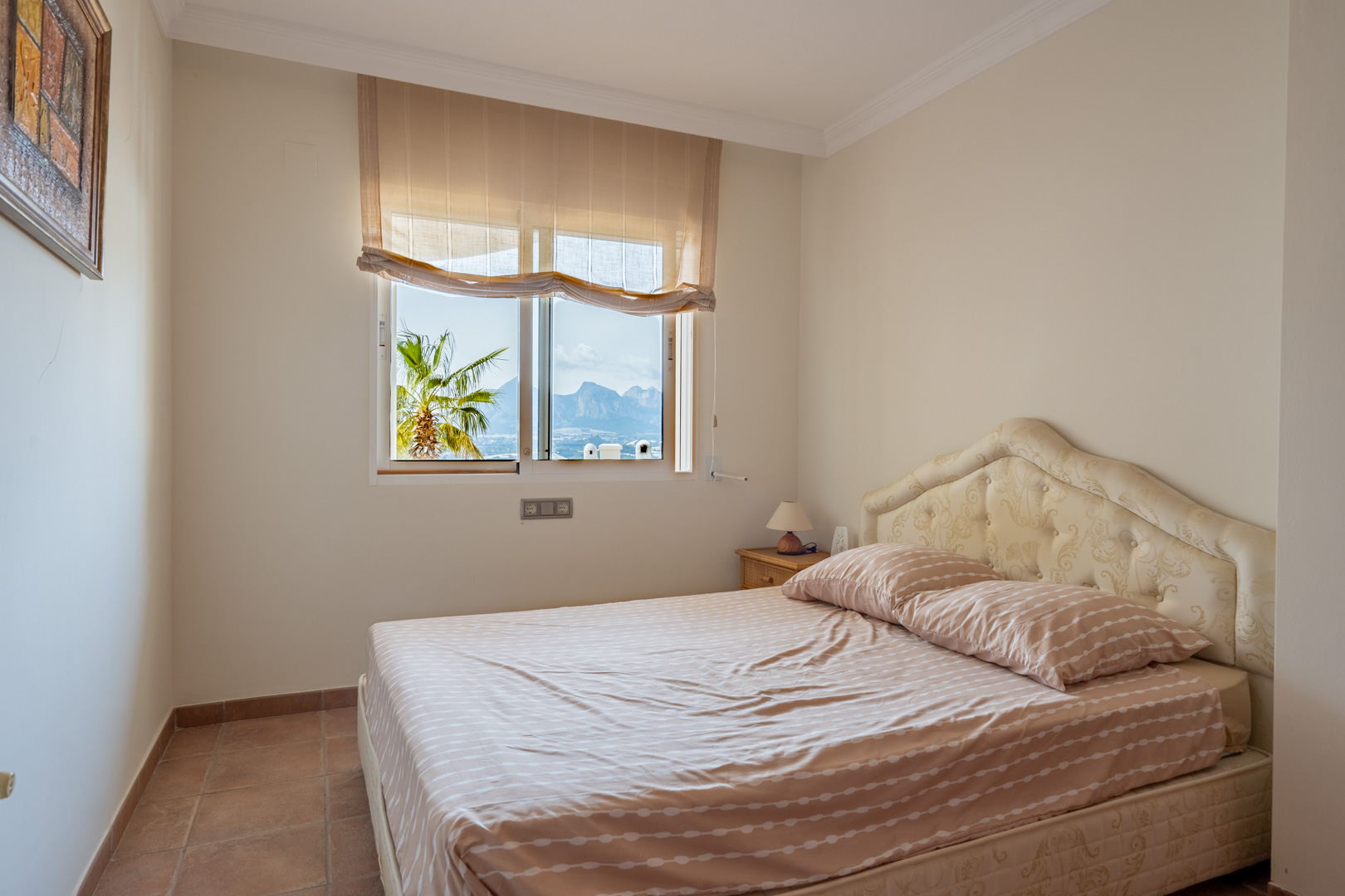 Dormitorio con suelos de gres y cama doble con cabecero tapizado. La ventana ofrece vistas directas a las montañas de Altea y a una palmera exterior.