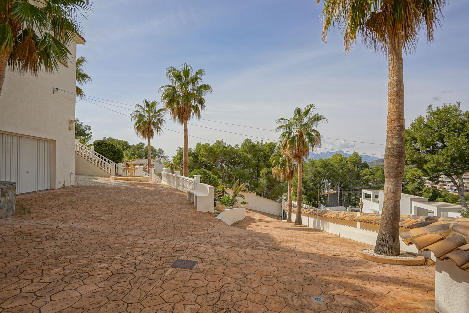 Entrada de la villa con amplio camino pavimentado en hormigón impreso, palmeras maduras y vistas a las montañas. Incluye acceso al garaje y balaustradas blancas.