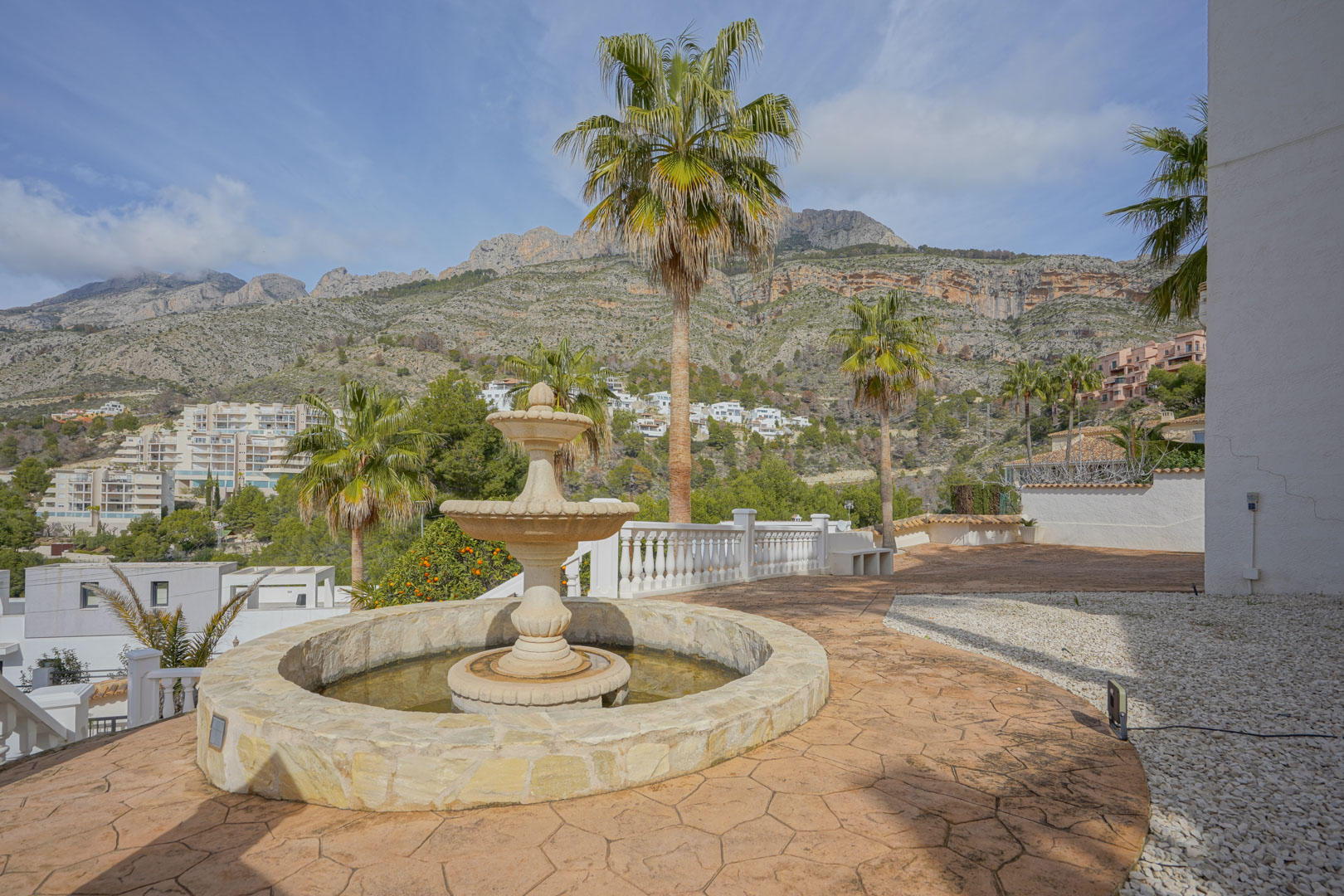 Terraza exterior con fuente de piedra tallada y suelos de hormigón impreso, con vistas panorámicas a la Sierra de Bernia y palmeras integradas.