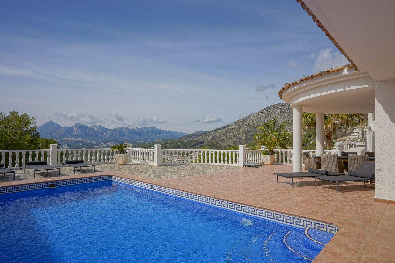 Terraza principal con piscina de mosaico azul, zona de solárium y porche cubierto. Ofrece vistas panorámicas a las montañas de Puig Campana y Sierra Aitana.