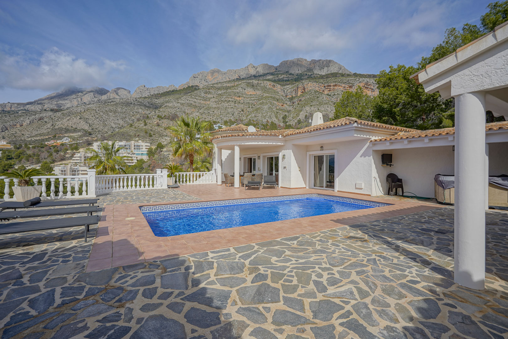 Terraza principal con piscina rectangular, borde de mosaico y vistas a la Sierra de Bernia. El espacio cuenta con pavimentos de piedra natural, baldosas de terracota y balaustrada blanca.