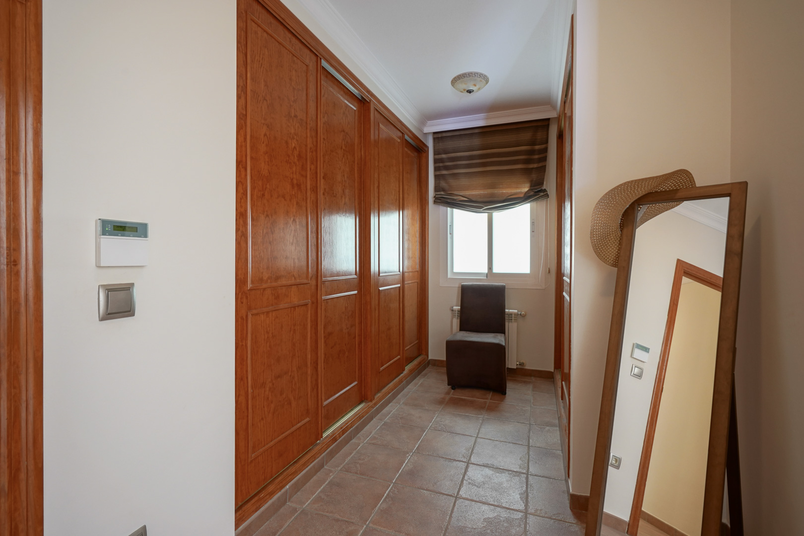 Vestidor con amplios armarios empotrados de madera, suelo de baldosas de terracota y ventana que aporta luz natural. Incluye espejo de cuerpo entero y silla auxiliar.