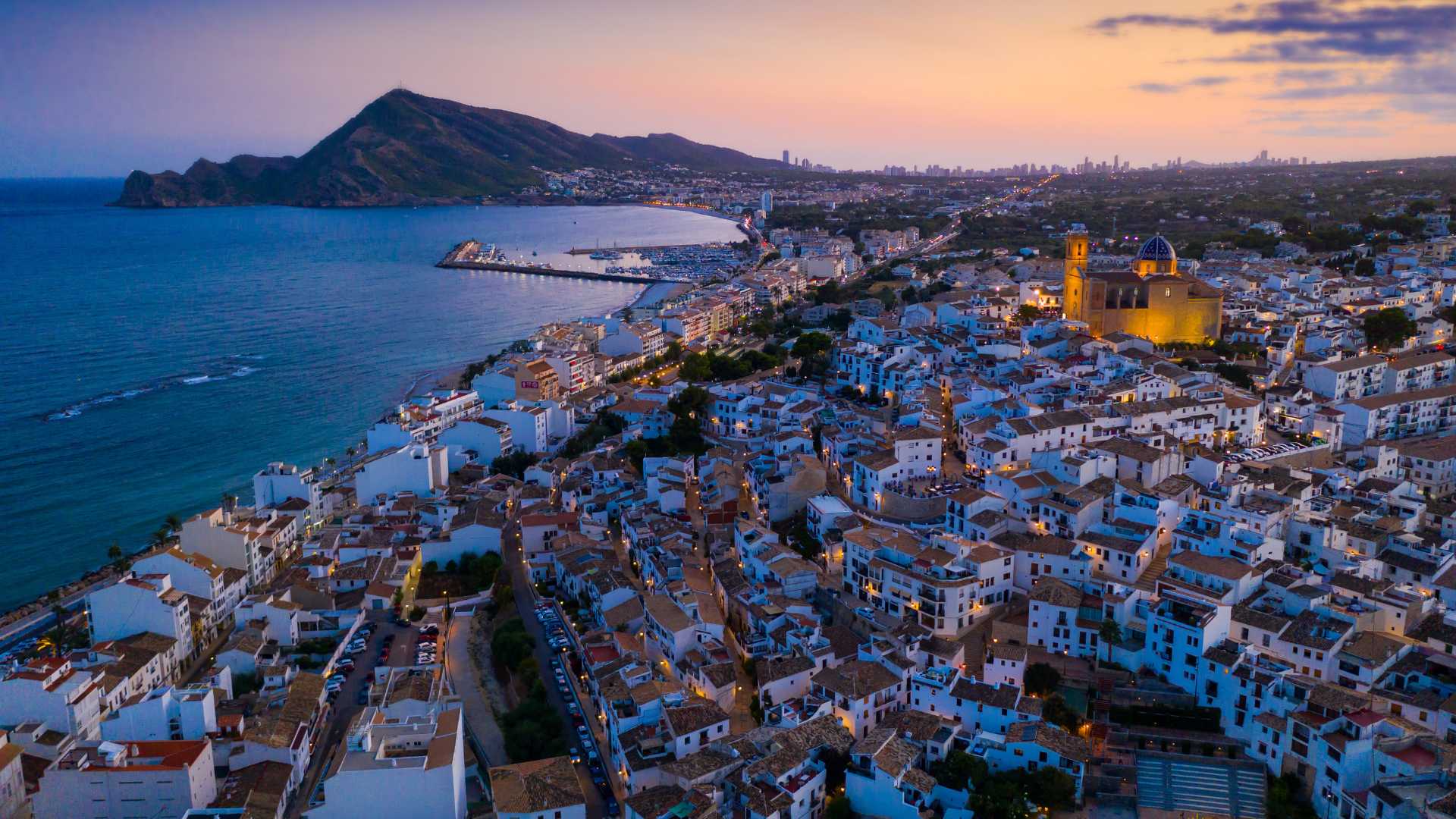 Vista aérea del casco antiguo de Altea al atardecer, destacando la iglesia con su cúpula azul, las casas blancas tradicionales y la silueta de la costa mediterránea.
