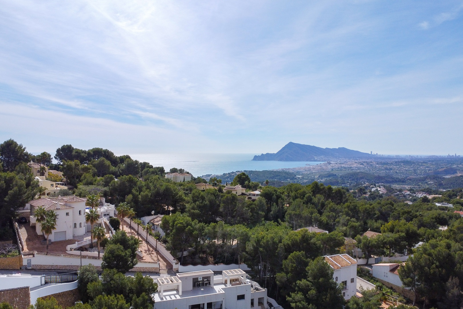 Vista aérea panorámica que muestra la densa vegetación de pinos, las villas blancas circundantes y el mar Mediterráneo con el perfil de Benidorm al fondo.