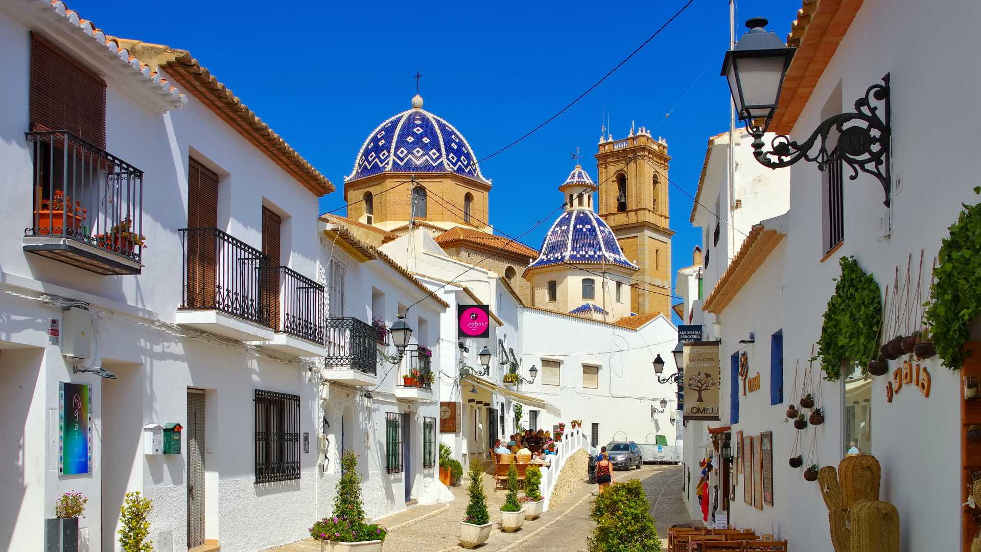 Vista del casco antiguo de Altea con sus calles empedradas, casas blancas y las icónicas cúpulas de azulejos azules y blancos de la iglesia principal bajo un cielo despejado.