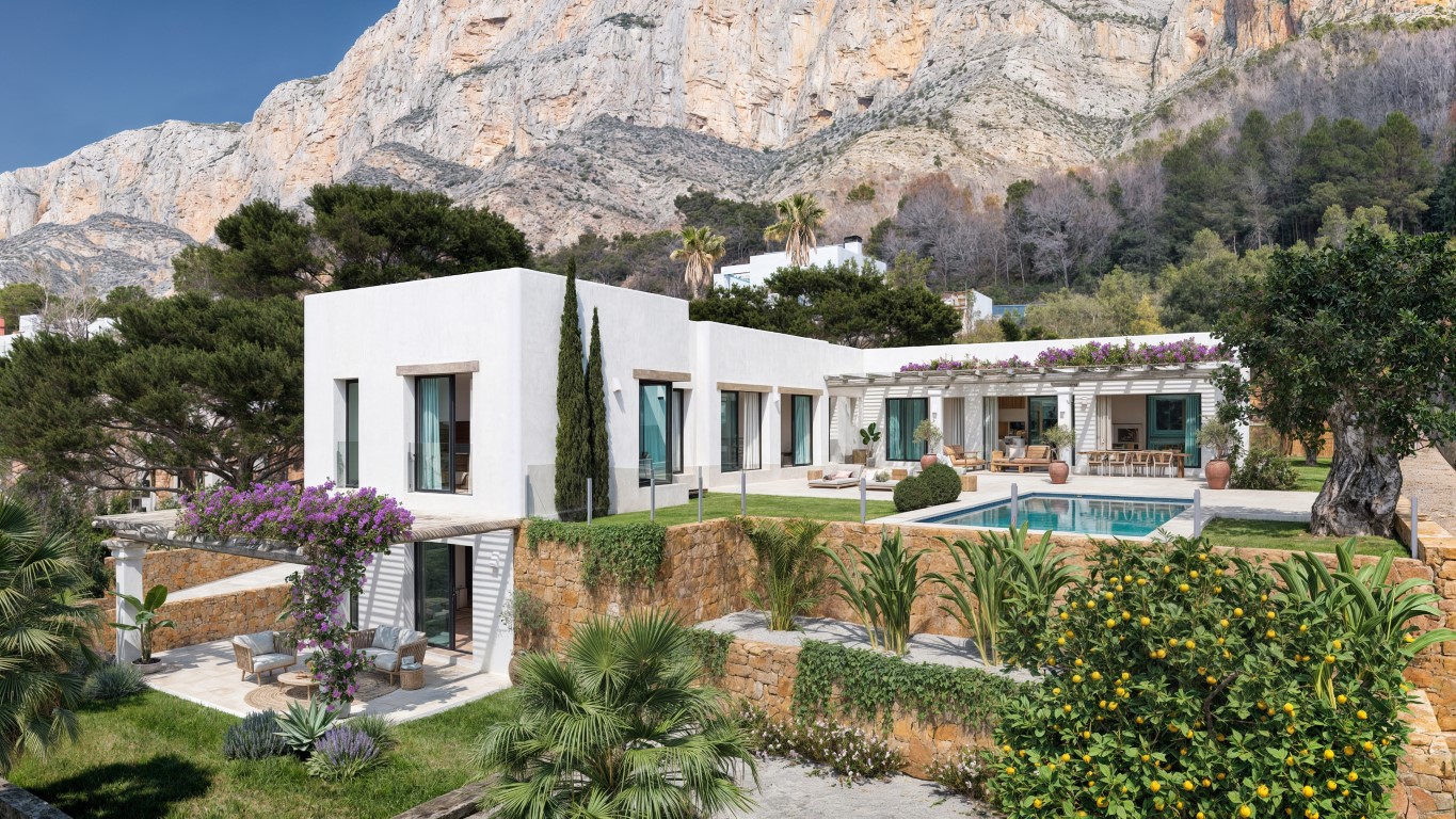 Vista exterior de la villa con fachada blanca, piscina privada y muros de piedra natural, situada bajo los impresionantes acantilados del Montgó en Jávea.