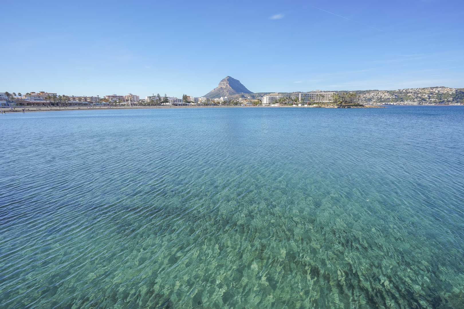 Vista panorámica de la bahía de Jávea con aguas cristalinas, mostrando el perfil del Montgó y la línea costera urbana bajo un cielo despejado.