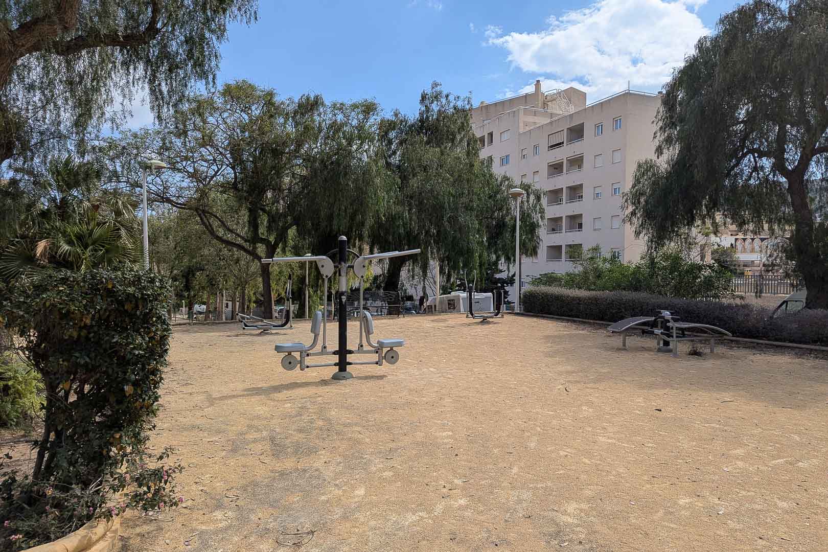 Área de gimnasio al aire libre con máquinas de ejercicio biosaludables, rodeada de vegetación autóctona y árboles maduros en un entorno residencial tranquilo.