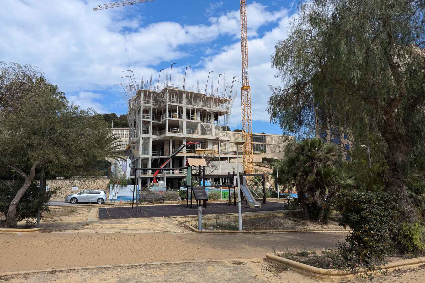 Estado actual de las obras en La Manzanera, Calpe, mostrando la estructura de hormigón armado y grúas de construcción junto a una zona de juegos infantiles.