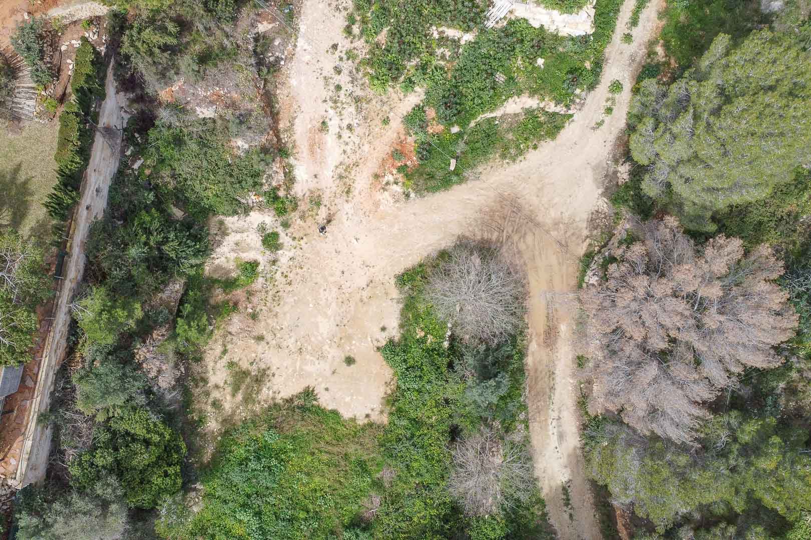 Vista aérea de la parcela en la urbanización La Corona, mostrando el terreno natural con vegetación autóctona y caminos de acceso antes del inicio de la construcción.
