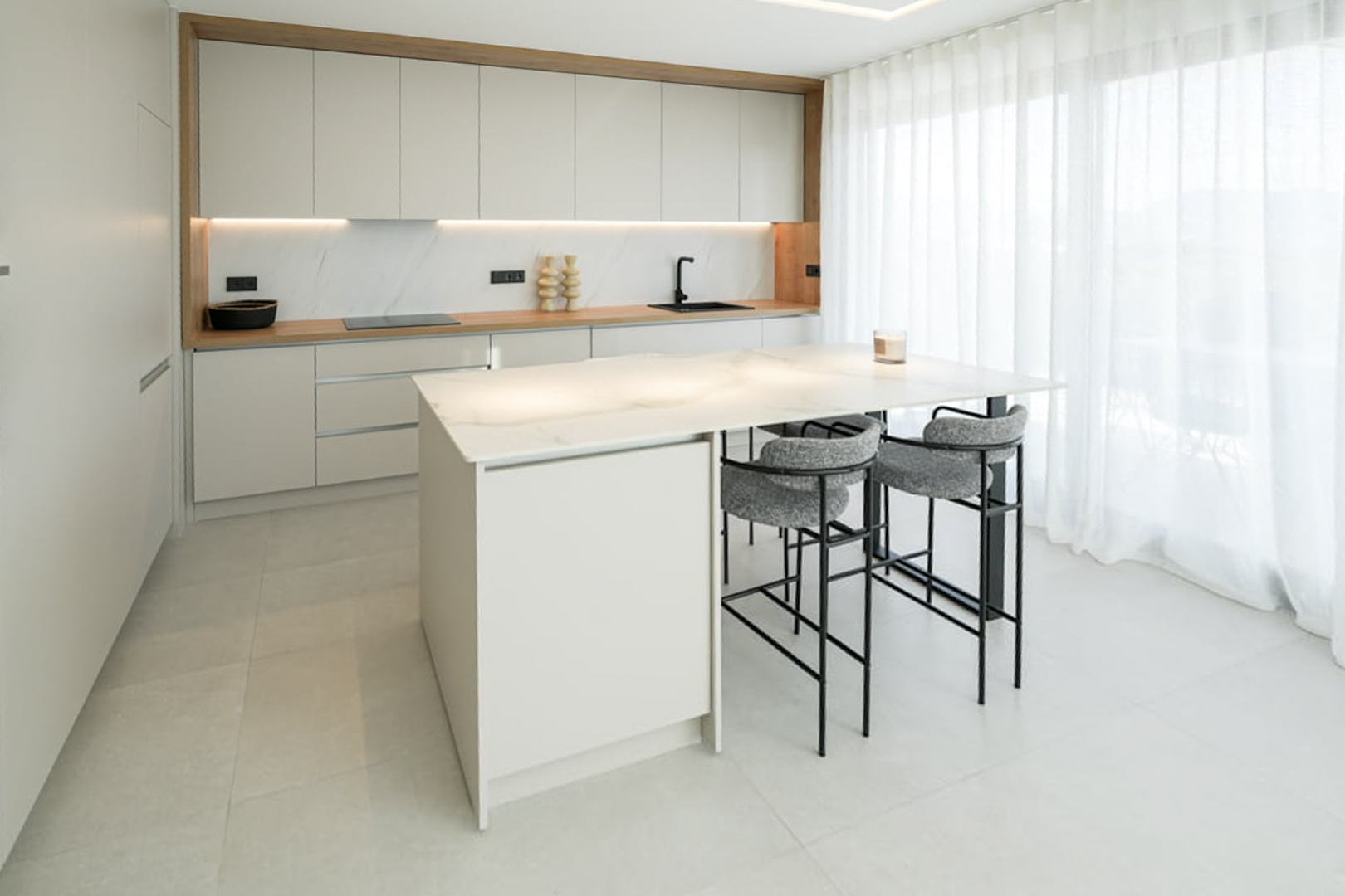 Cocina moderna con isla de mármol, taburetes grises y gabinetes blancos bajo luz natural.