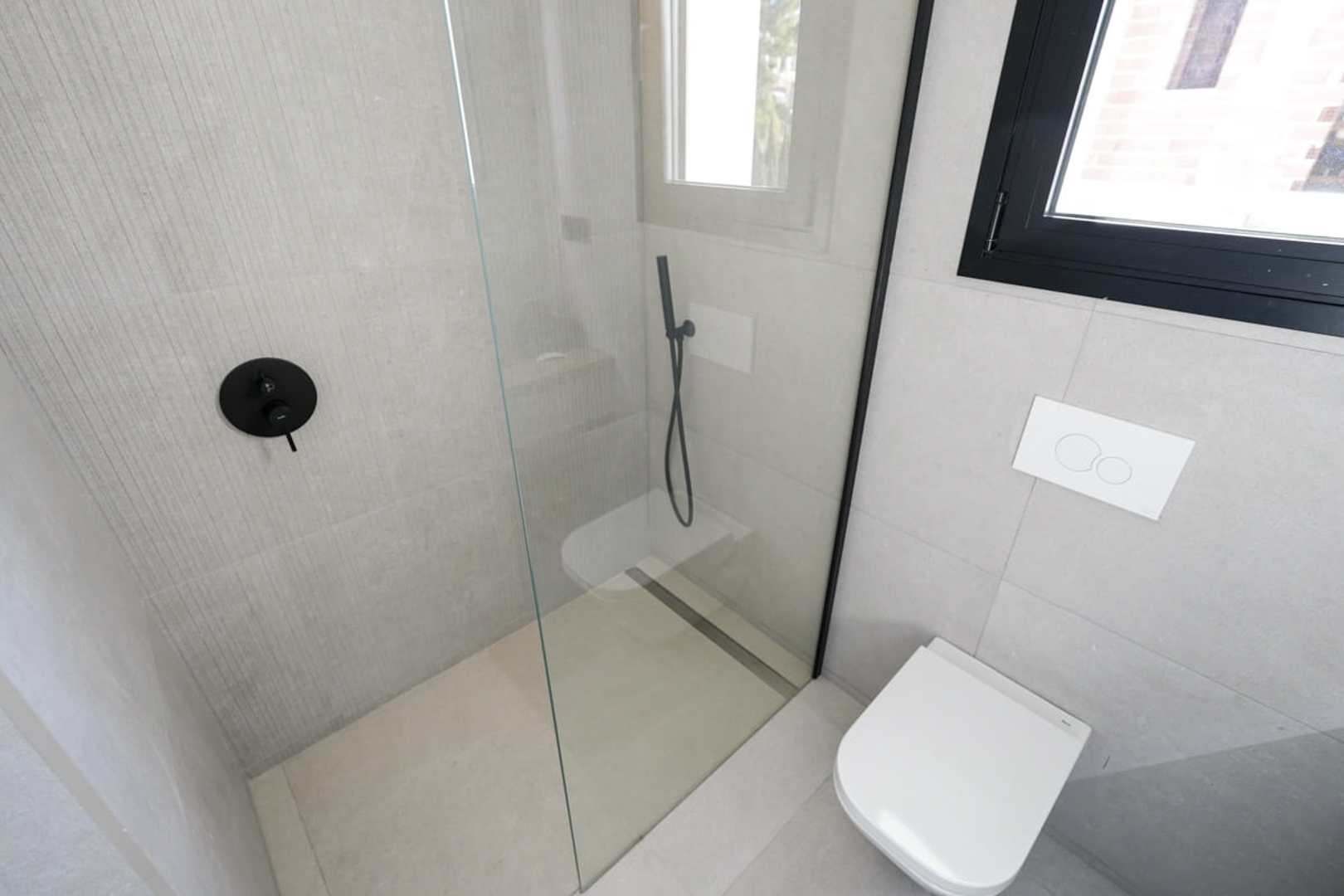 Detalle de baño moderno con ducha de vidrio, grifería negra y azulejos grises texturizados.