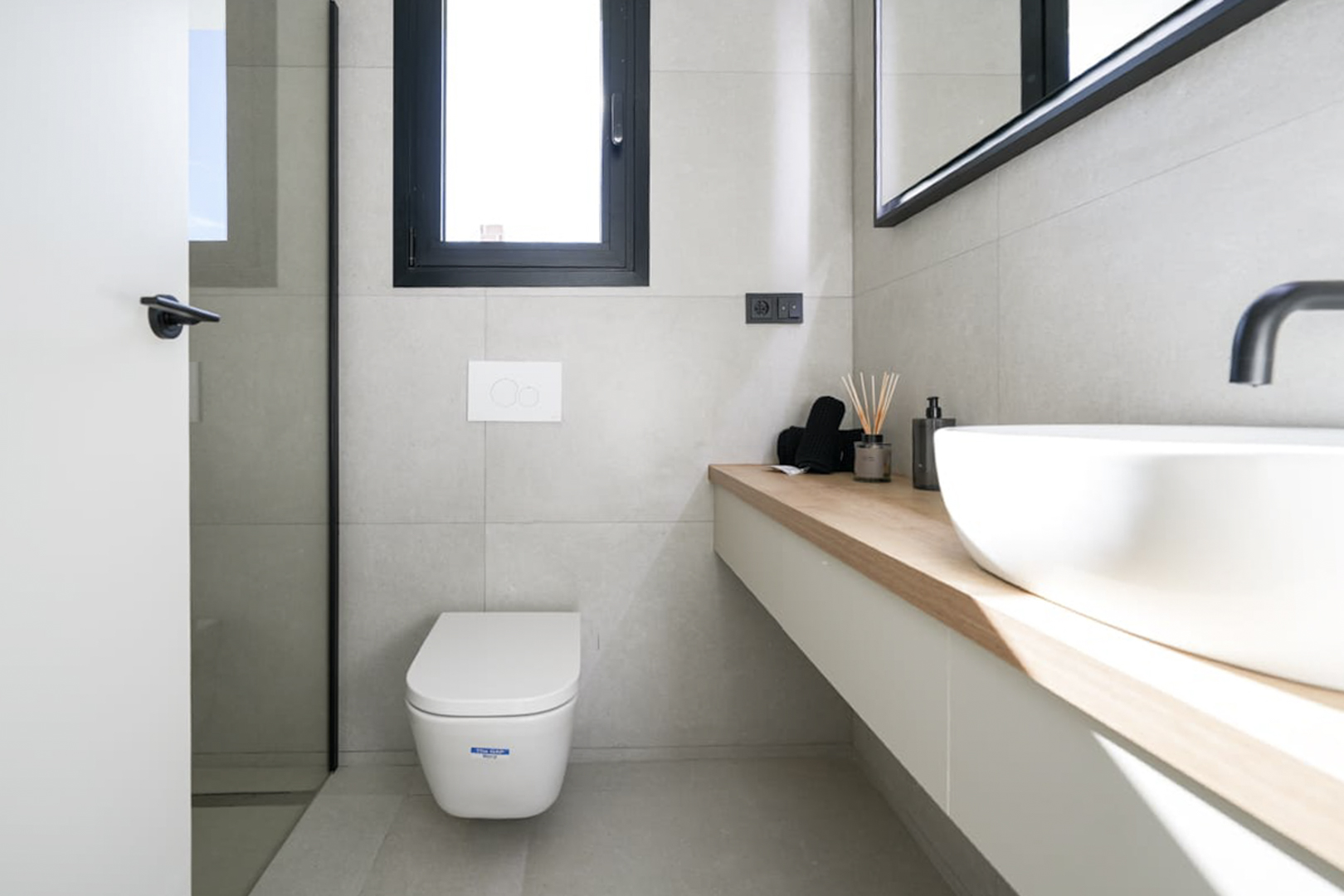 Detalle de baño moderno con encimera de madera clara, lavabo blanco y azulejos grises neutros.