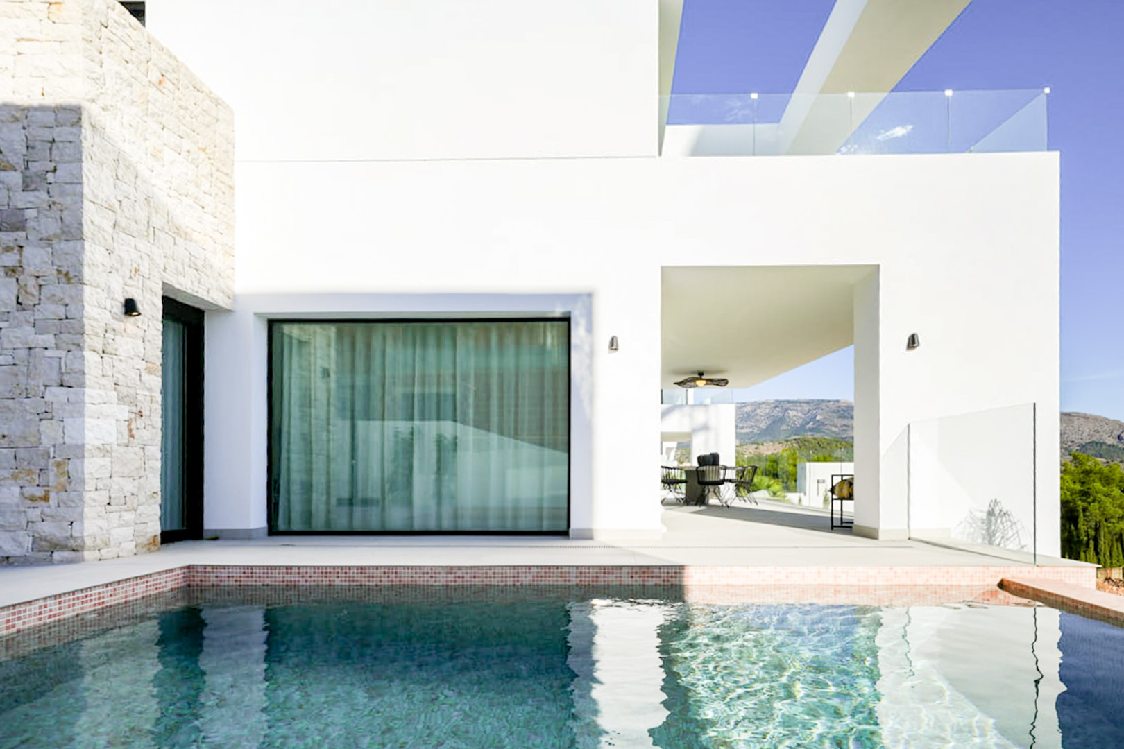 Vista de la piscina con borde de mosaico, la fachada de piedra y la terraza cubierta con comedor exterior.