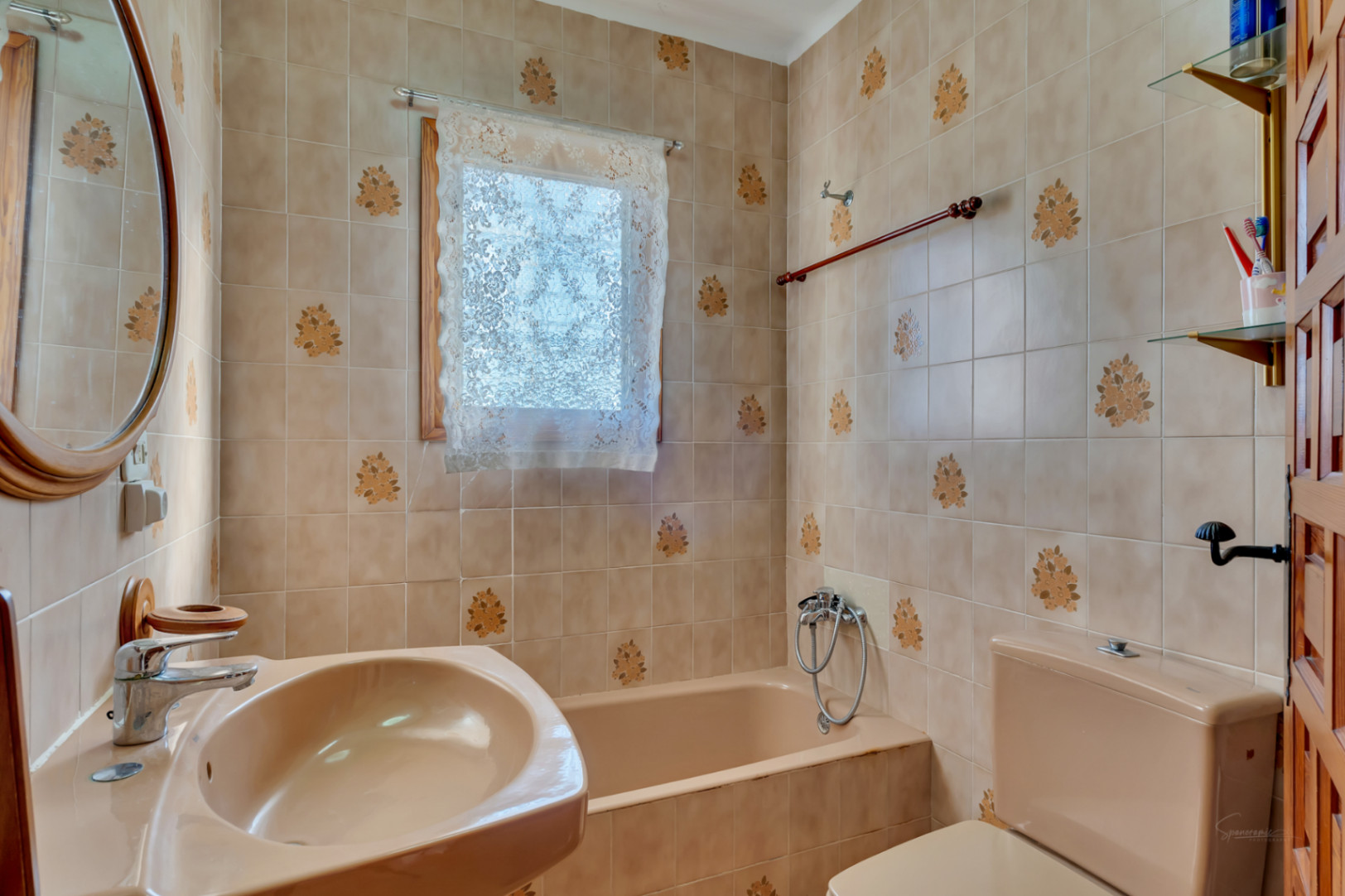 Baño de estilo tradicional con azulejos decorados con motivos florales, bañera encastrada y lavabo en tonos beige, complementado con un espejo circular de madera.