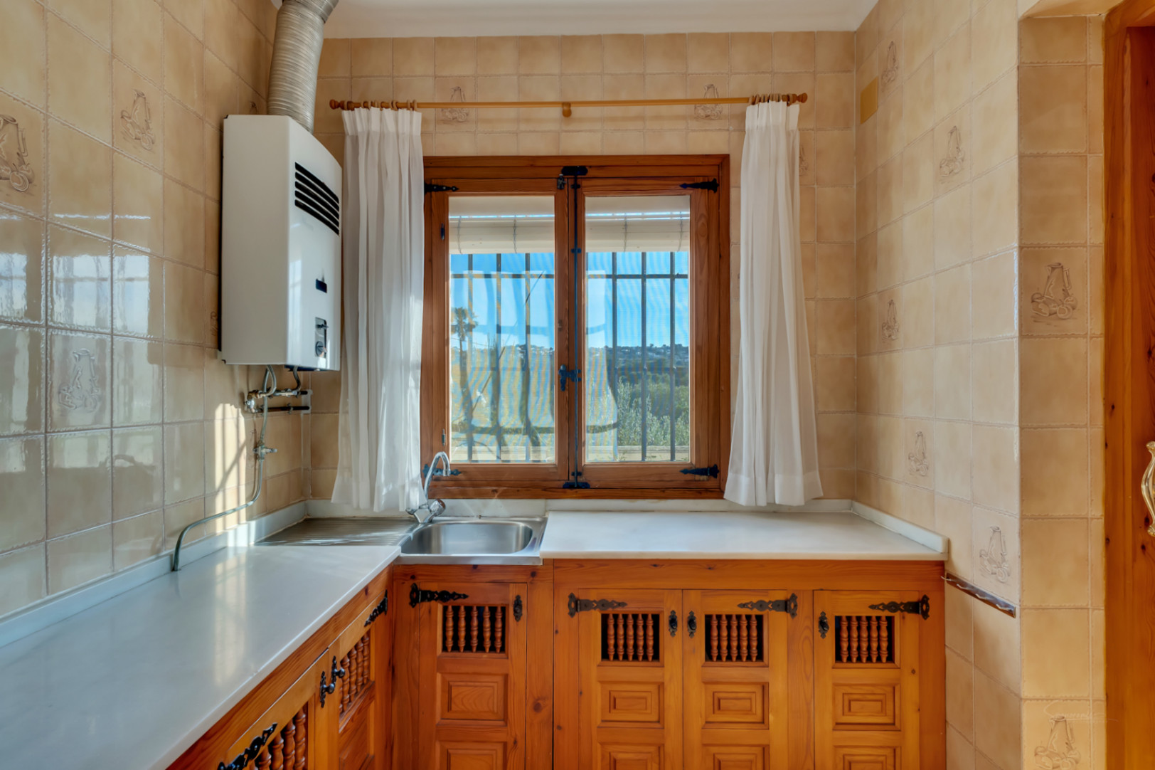 Cocina de estilo tradicional con armarios de madera tallada, encimera blanca y ventanal de madera que aporta luz natural y vistas despejadas.