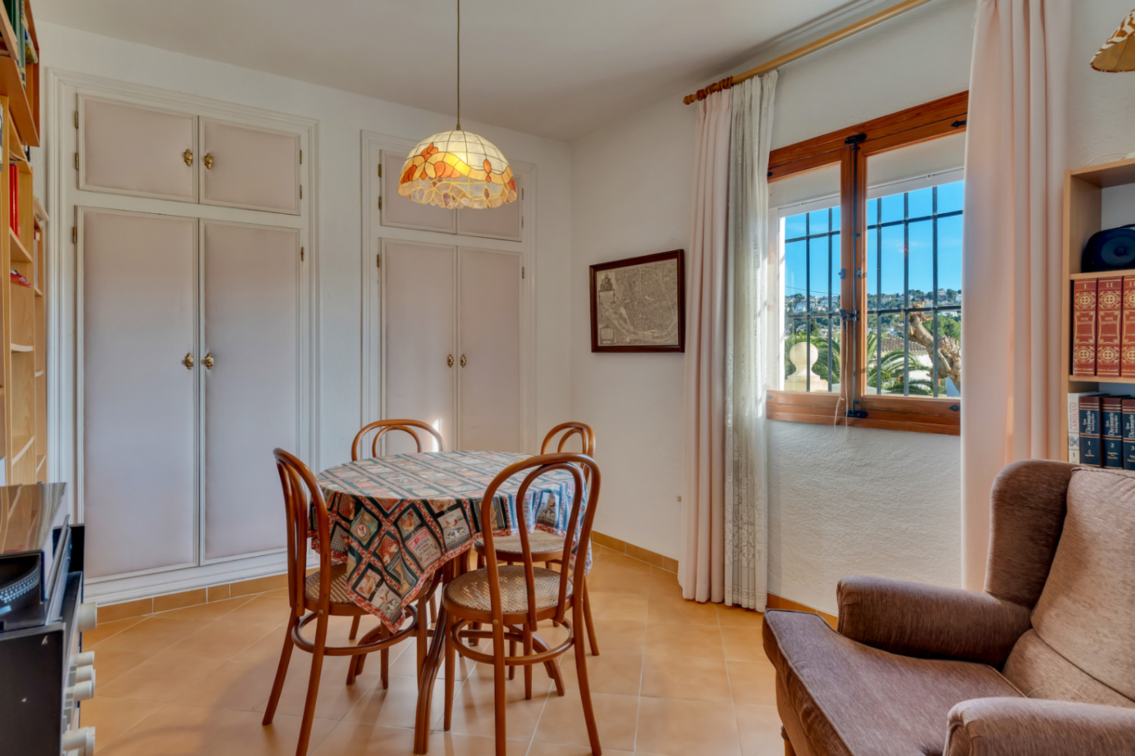 Comedor luminoso con armarios empotrados blancos, mesa redonda y sillas de madera. La ventana con marco de madera ofrece luz natural y vistas al entorno residencial de Jávea.