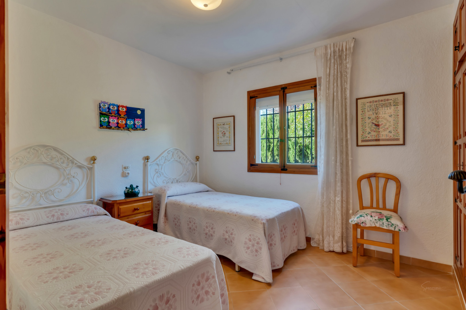 Dormitorio luminoso con dos camas individuales, cabeceros de forja blanca, suelos de terracota y ventana de madera con luz natural.