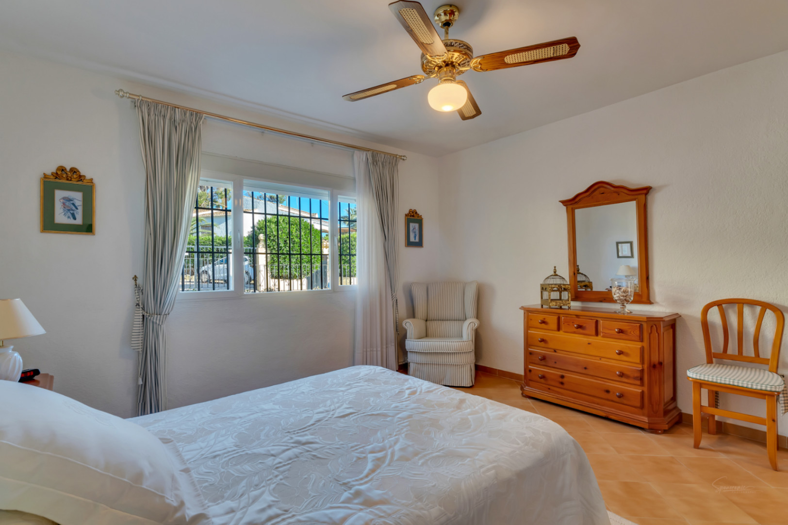 Dormitorio luminoso con suelos de gres, mobiliario de madera clásica, ventilador de techo y ventana con vistas al jardín exterior.