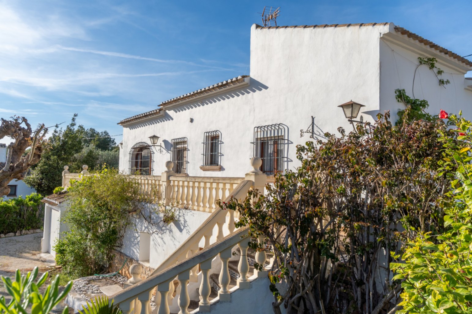 Fachada blanca de estilo mediterráneo con escalera exterior de balaustrada, ventanas protegidas por rejas de hierro y vegetación consolidada.