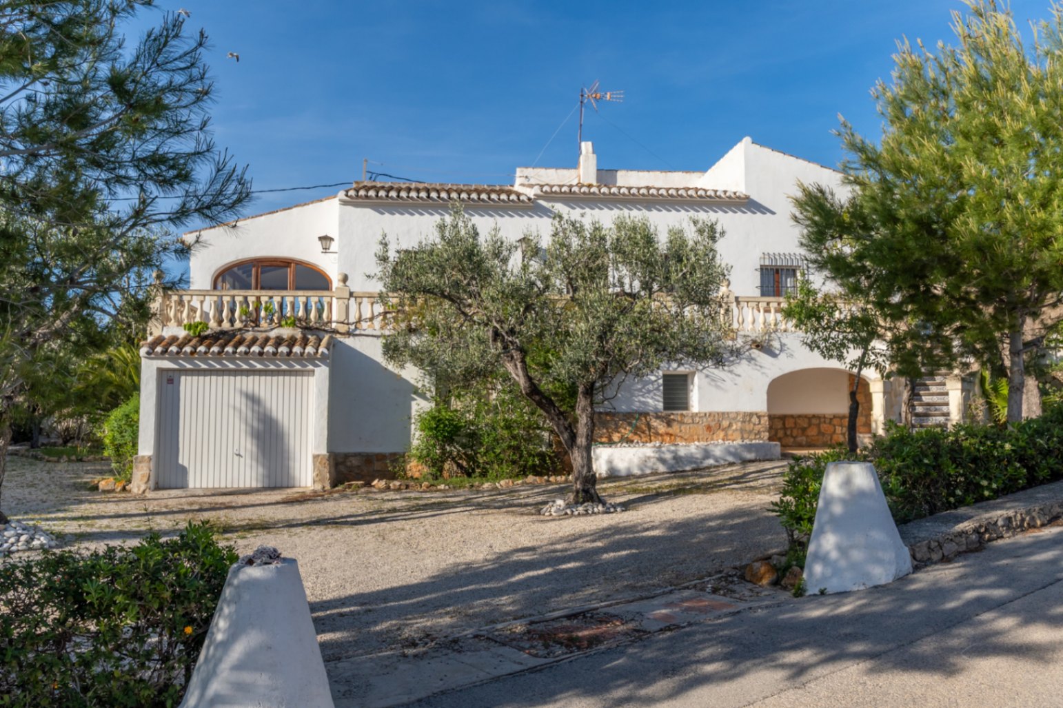 Fachada de la villa con arquitectura tradicional mediterránea, garaje privado, terraza superior con balaustrada y jardín con olivos centenarios sobre zona de grava.