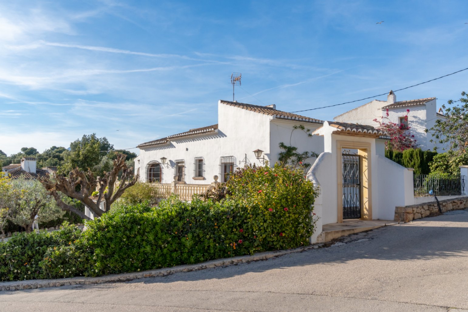 Fachada exterior de estilo mediterráneo con muros blancos, carpintería de hierro forjado y entrada peatonal enmarcada en piedra tosca tradicional.