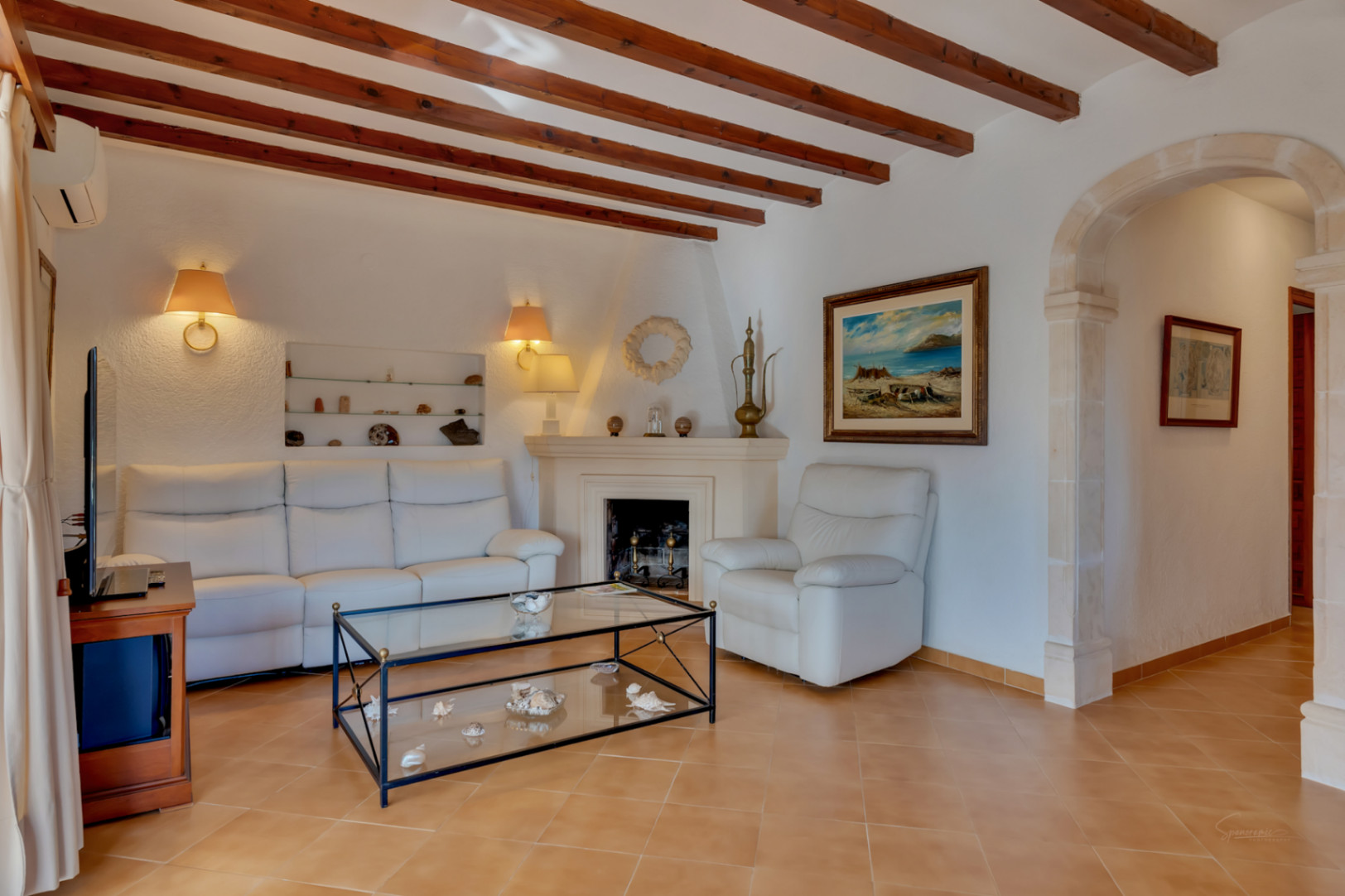 Salón con vigas de madera, chimenea clásica y un arco de piedra tosca tradicional. Mobiliario en tonos blancos sobre suelos cerámicos y estantería de obra integrada.