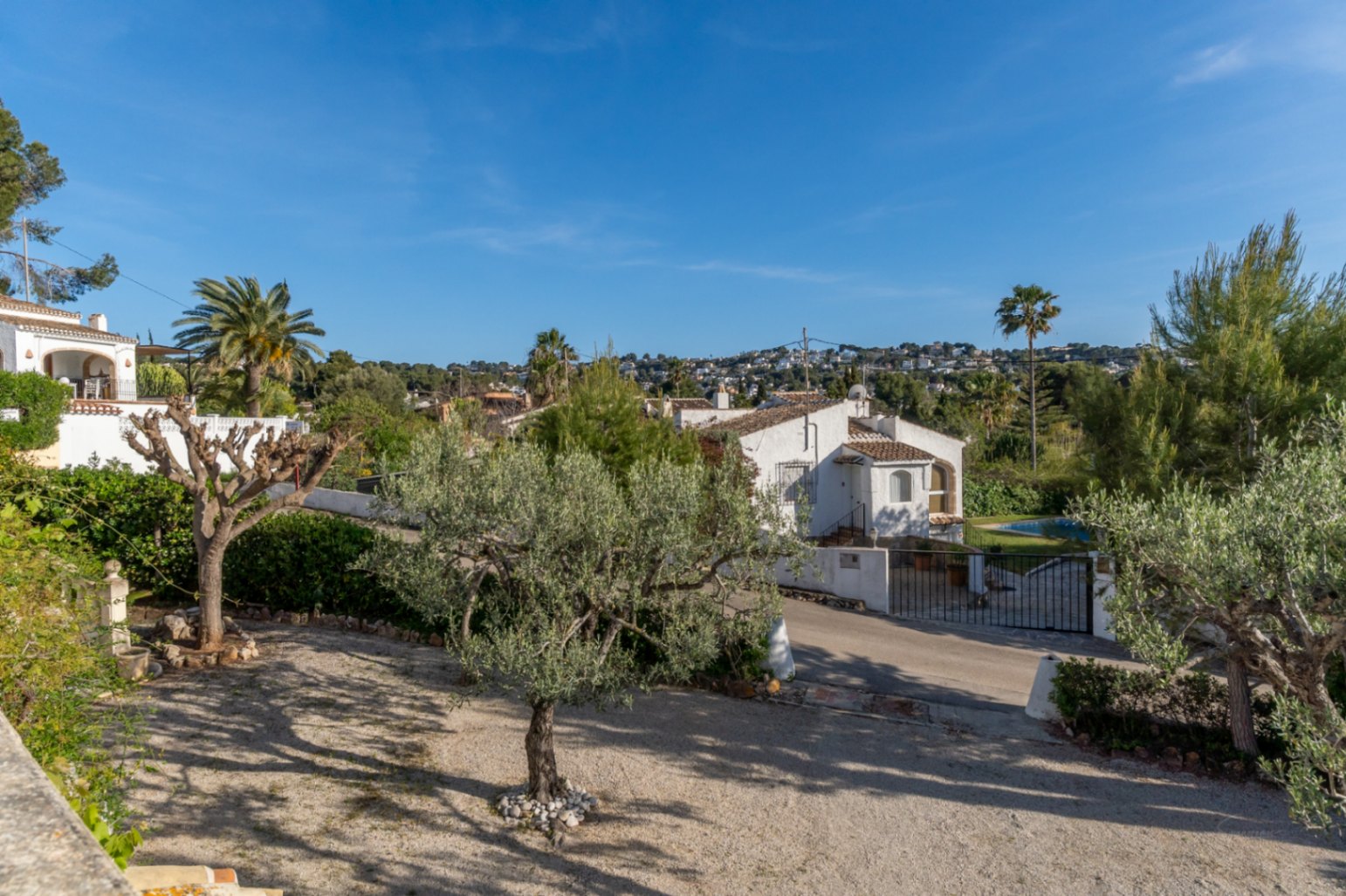 Vista exterior elevada que muestra el jardín de grava con olivos y la arquitectura tradicional de la villa bajo un cielo despejado en Jávea.