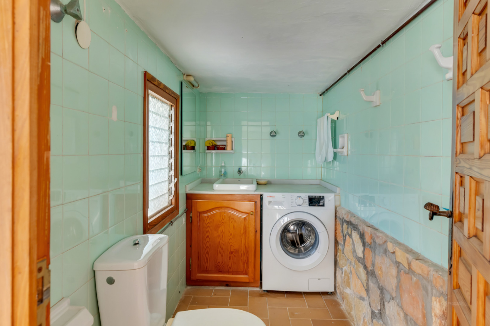 Zona de lavandería y aseo con azulejos verde menta, pared de piedra natural, lavadora integrada y mueble de madera bajo el lavabo.