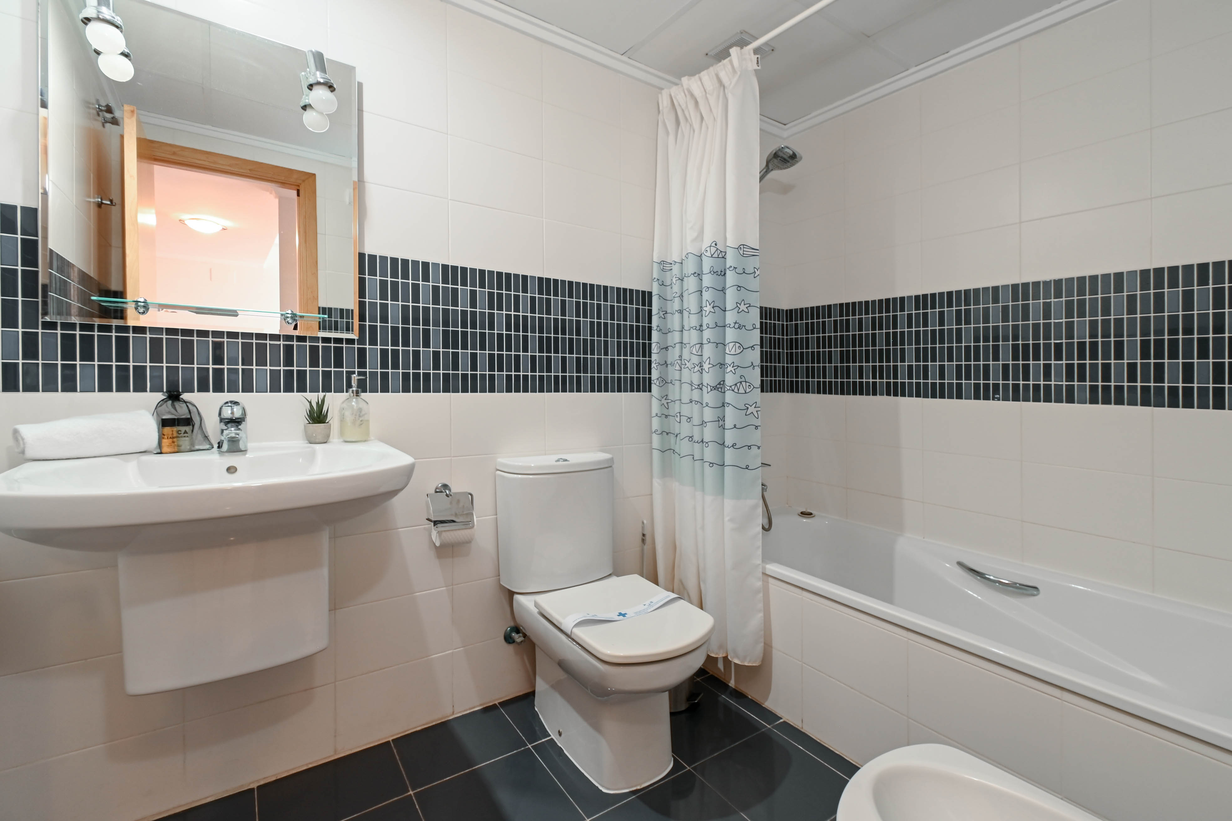 Baño funcional con azulejos blancos y franja de mosaico oscuro, equipado con bañera, lavabo suspendido y espejo con iluminación frontal.
