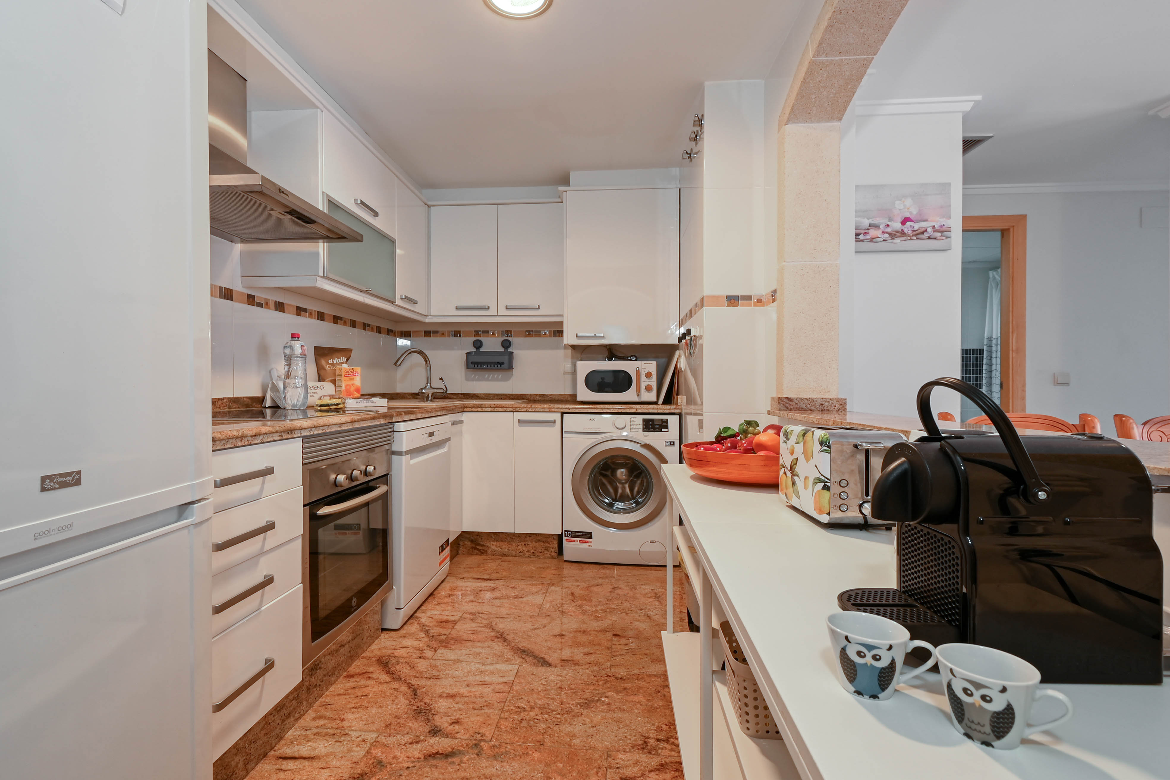 Cocina equipada con armarios blancos, encimeras de granito y electrodomésticos integrados como lavadora y lavavajillas, comunicada mediante un arco de piedra.