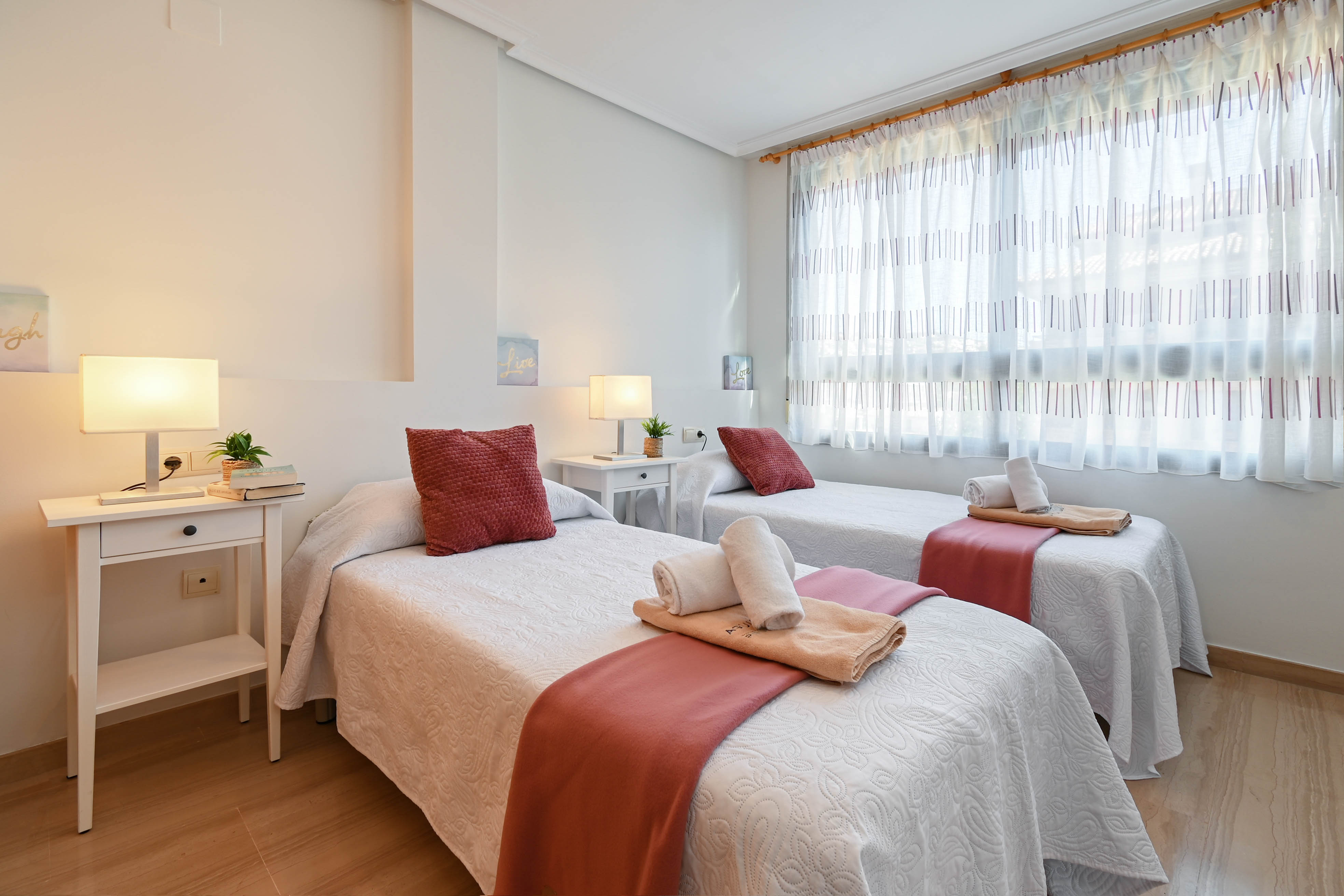 Dormitorio luminoso con dos camas individuales, mesitas de noche blancas y suelos de madera clara. Dispone de un gran ventanal con cortinas traslúcidas y lámparas de diseño.