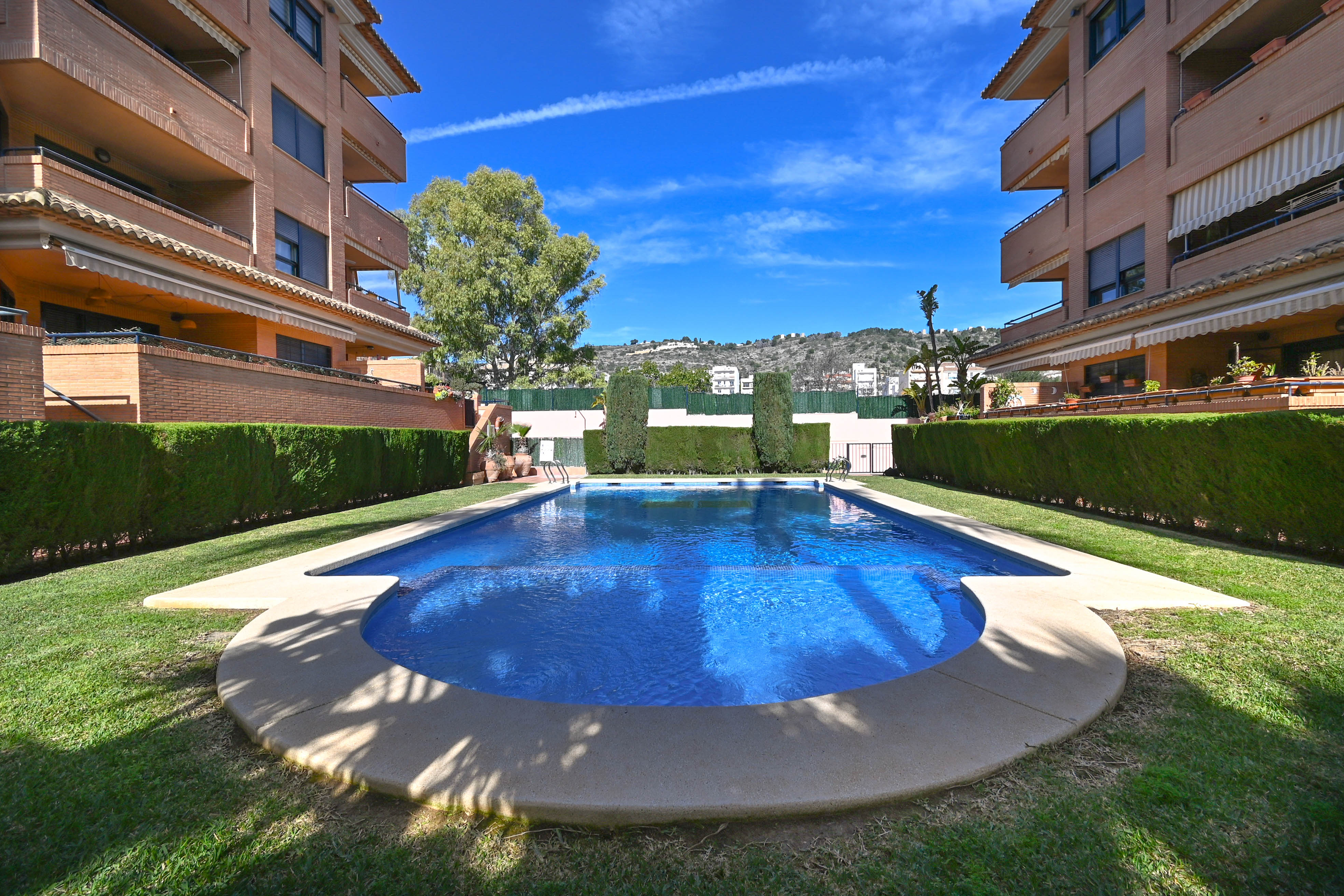 Piscina comunitaria con entrada semicircular, rodeada de césped y setos cuidados, situada entre edificios residenciales con vistas a las montañas.