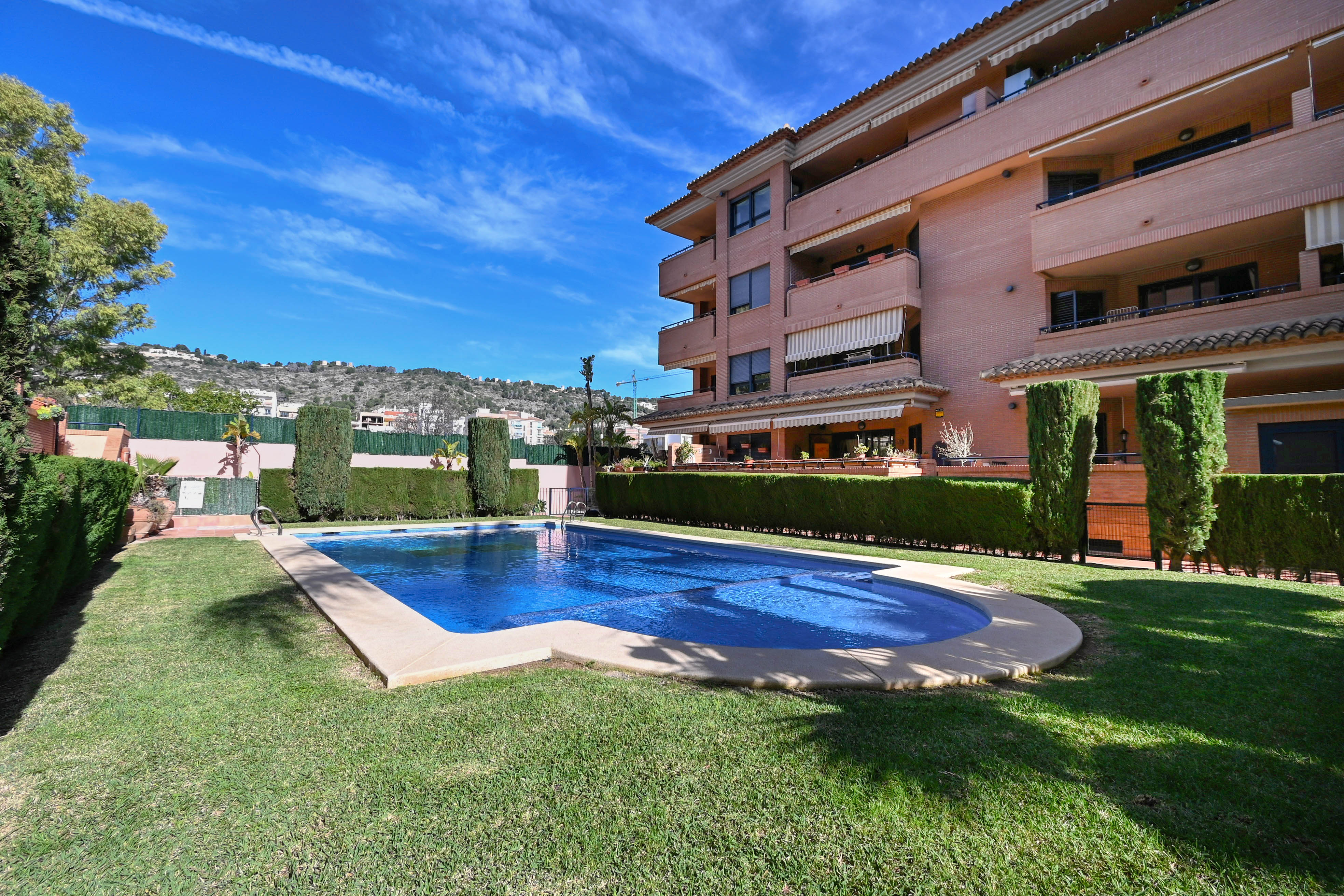 Piscina comunitaria rodeada de césped y setos altos, situada frente a un edificio de ladrillo visto bajo un cielo despejado en Jávea.